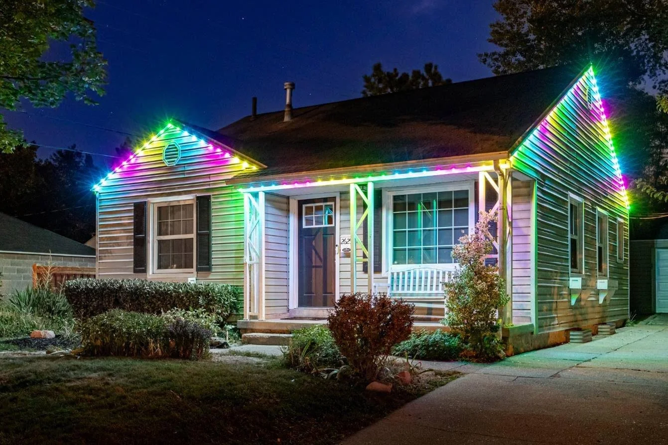 A house decorated with colorful neon lights outlining the roof, front porch, and sides, nighttime scene, with plants and bushes in front.