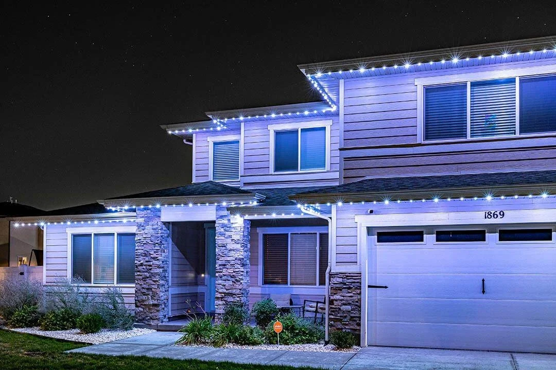 A two-story house decorated with white Christmas lights at night, with stone accents on the front facade and a white garage door.