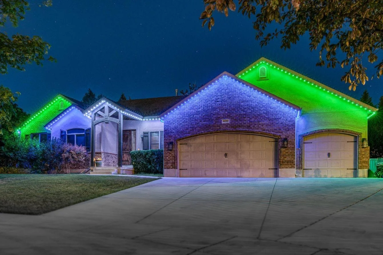 Nighttime exterior of a house decorated with colorful string lights, featuring a double garage and a front porch, with trees in the background.