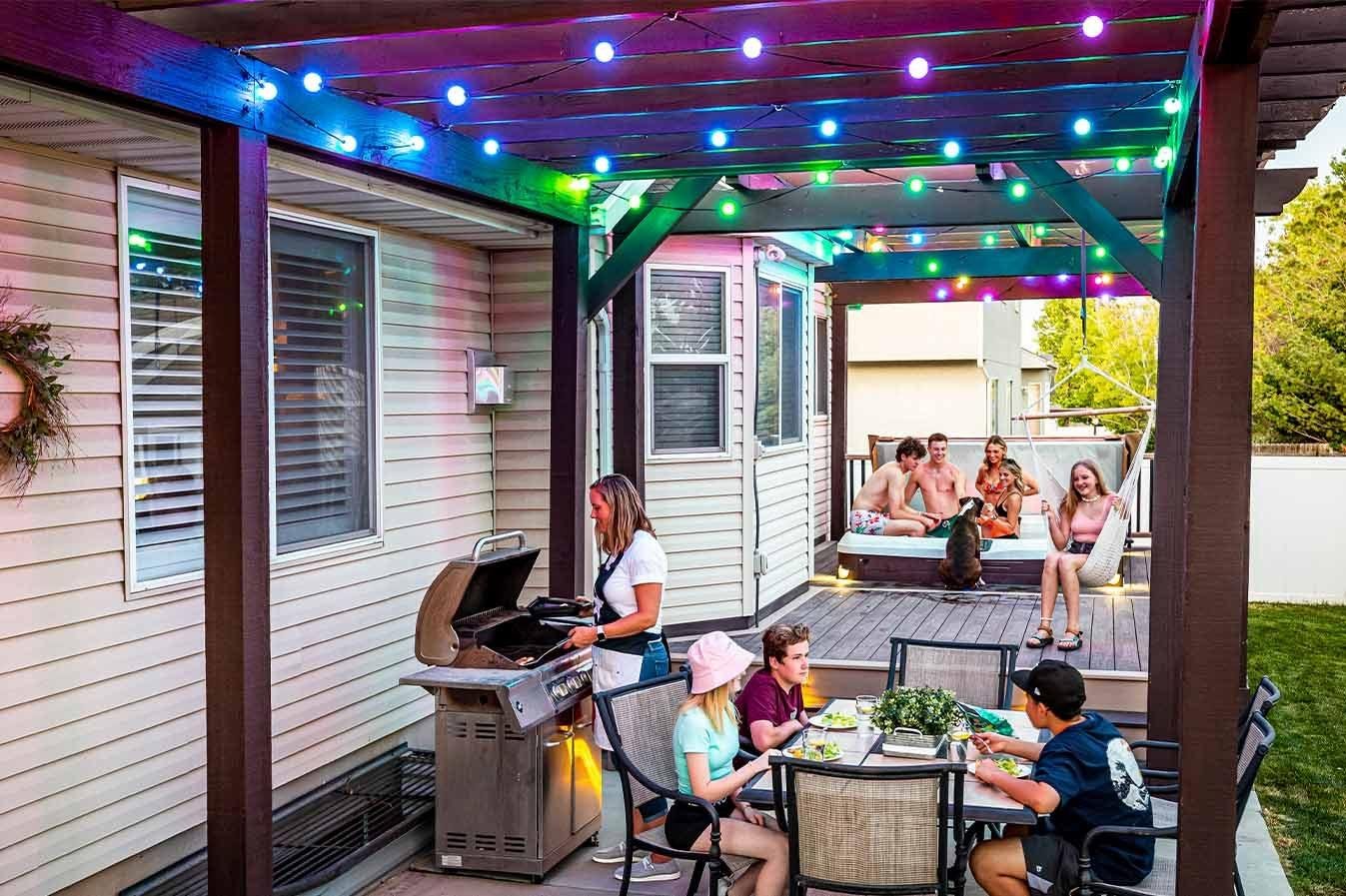 People enjoying a backyard gathering with string lights, a BBQ grill, and a deck with seating and a hot tub.