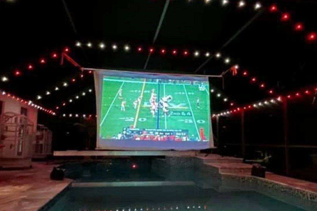 Outdoor pool area at night with a large projection screen displaying a football game, decorated with string lights overhead.