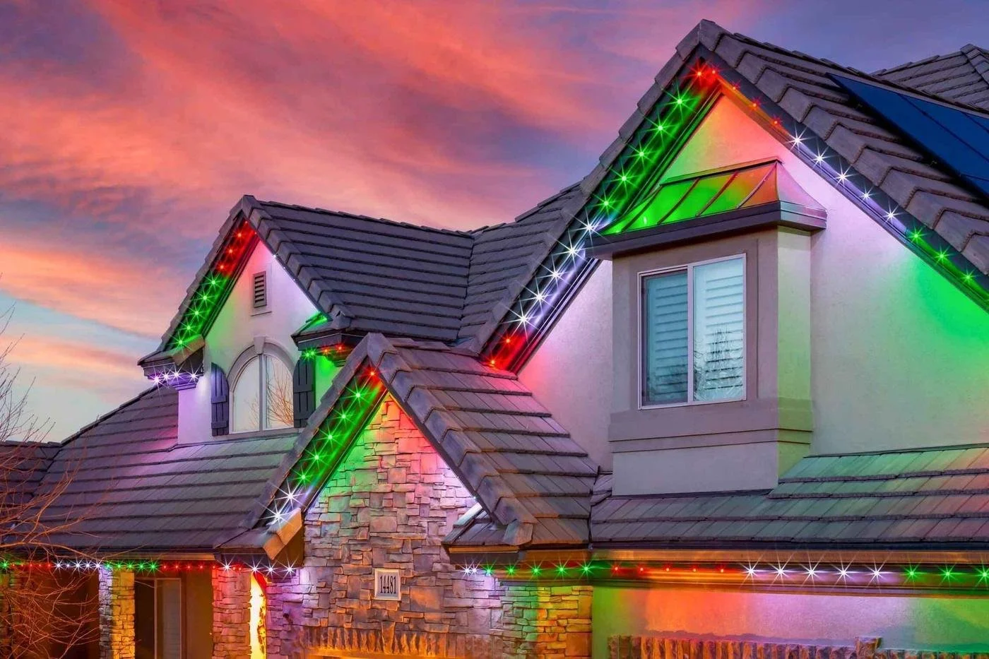 A house decorated with multicolored Christmas lights, including red, green, and purple, on the roof and around the windows, during sunset with a colorful sky.