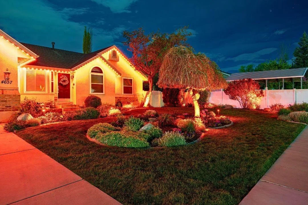 A house decorated with Christmas lights and wreath, with a well-maintained front yard featuring trees, shrubs, and a garden bed, illuminated at night.