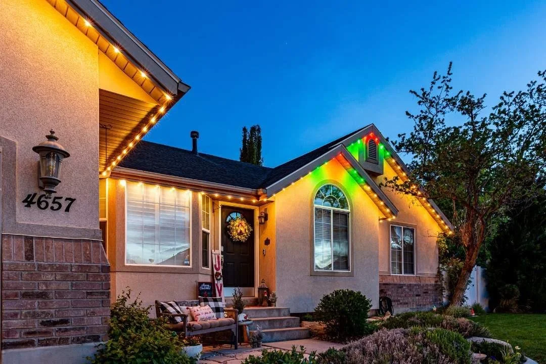 A house decorated with colorful Christmas lights on the roofline and a wreath on the front door, with a garden and trees in the yard during evening.