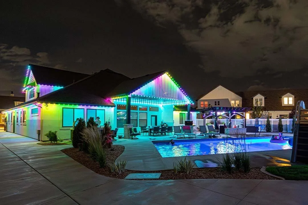 Nighttime view of a house with colorful string lights, a swimming pool with kids playing, lounge chairs, and a backyard patio area.