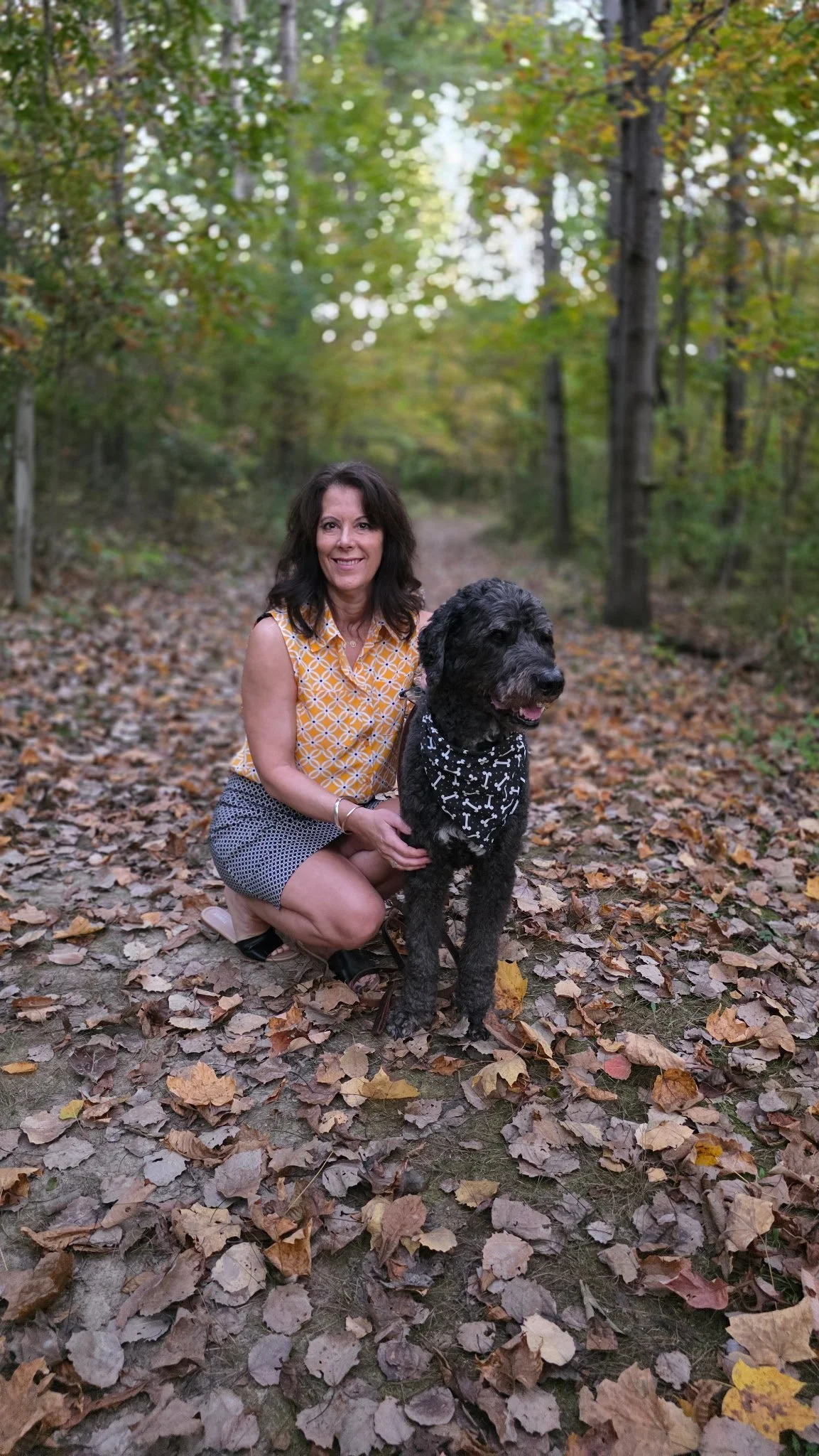 Woman kneeling beside a large black dog wearing a bone-print bandana on a leaf-covered forest trail