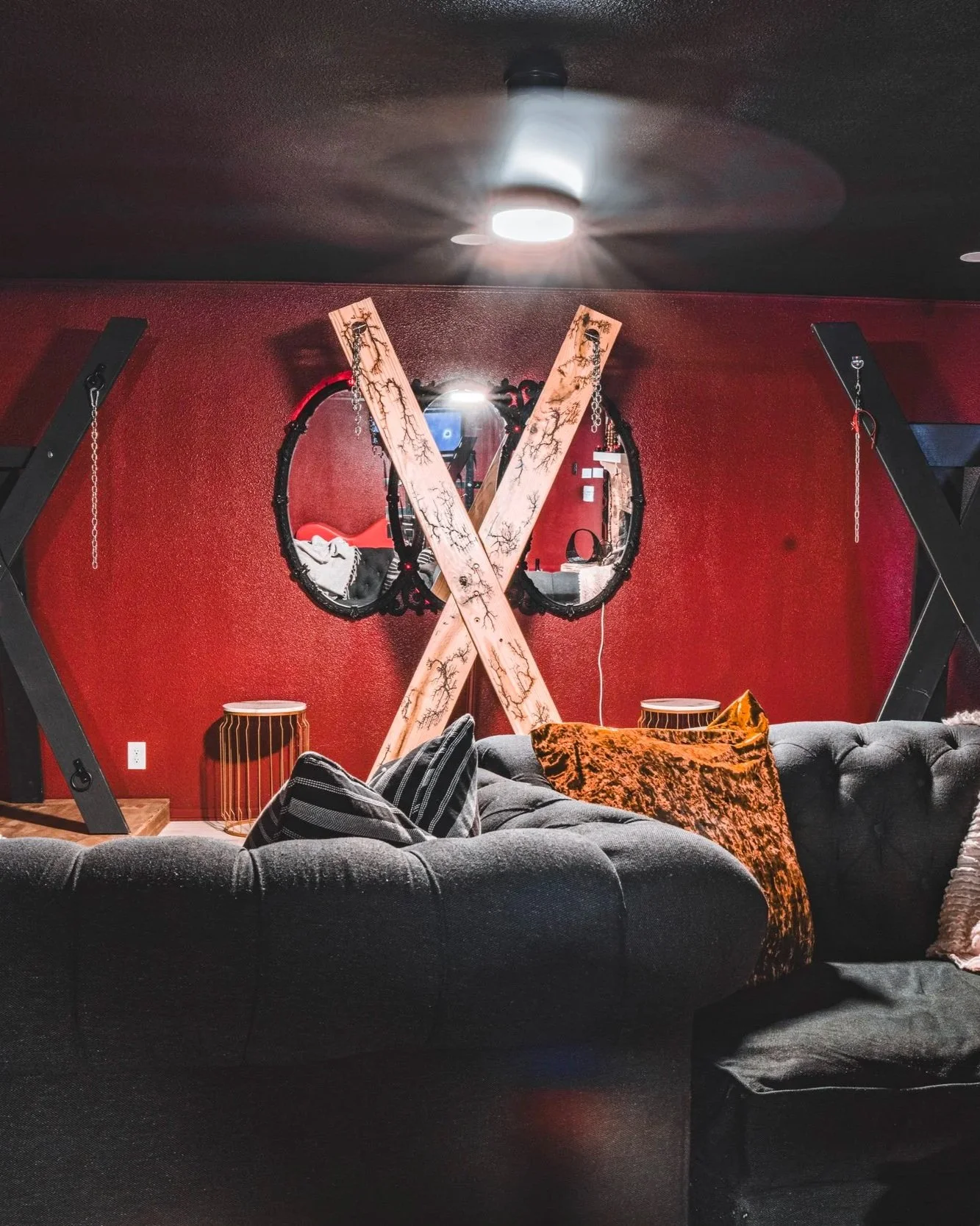 Interior of a room with red textured wall, ceiling fan, decorative mirrors, wooden criss-crossed beams, black workout equipment, side table, gray couch with striped pillow and orange patterned blanket.