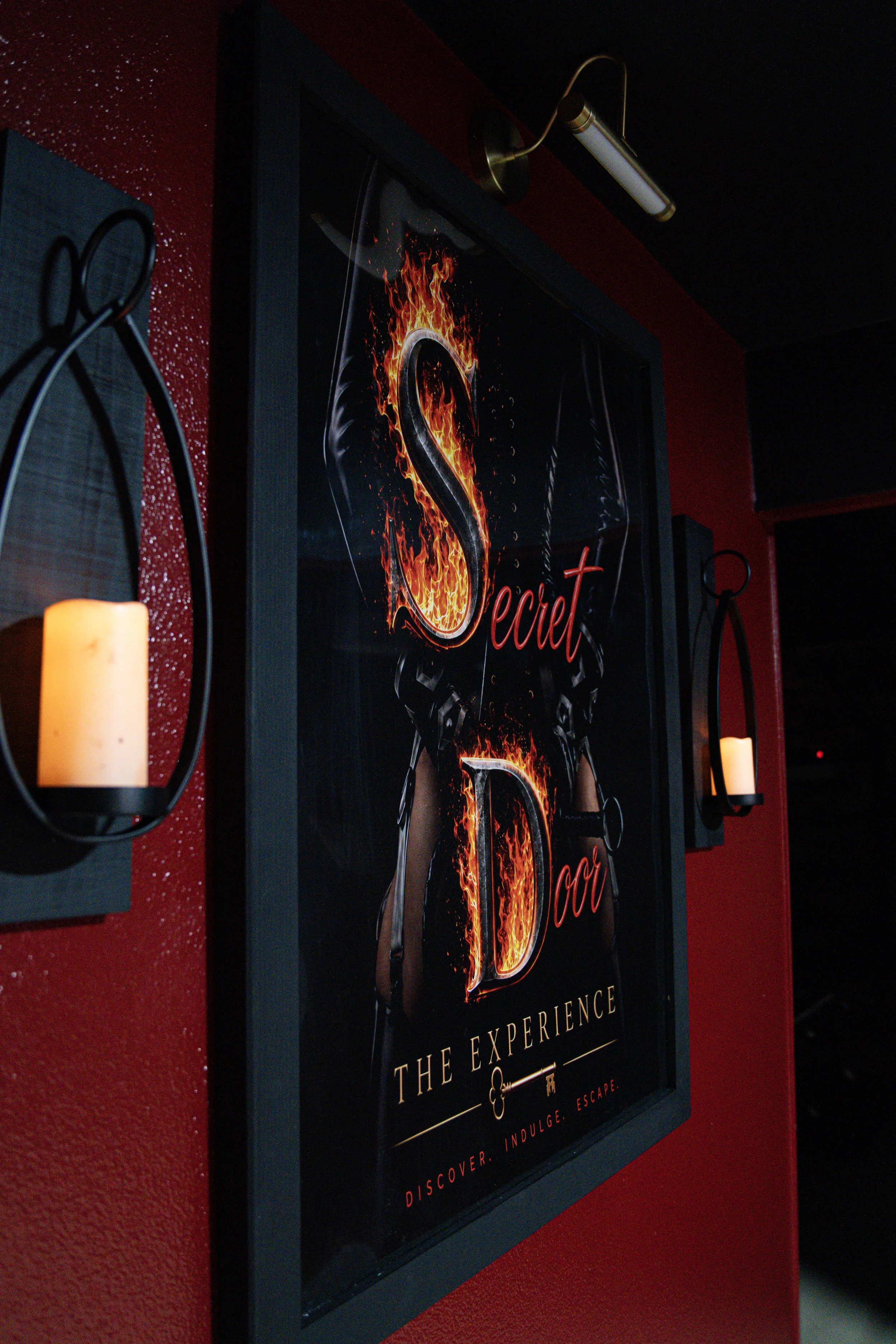 A black and red poster with the word "Secret" written in fiery letters, framed on a dark red wall, with candle holders on either side and a gold picture light above.