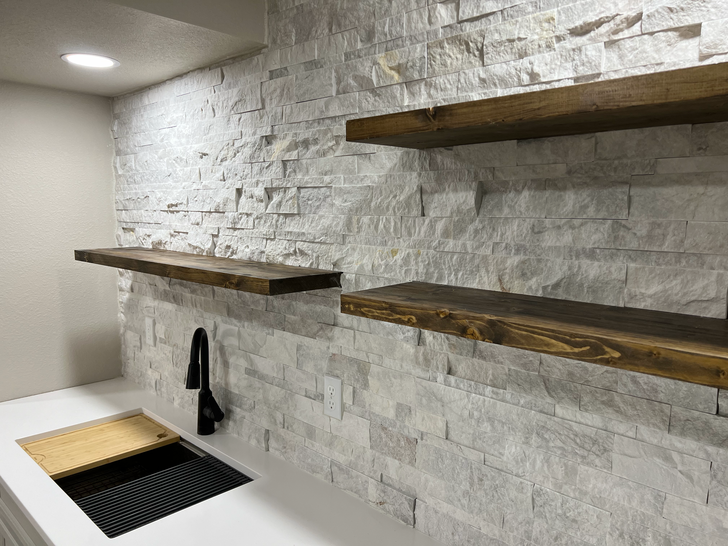 A kitchen with a white textured stone backsplash, a black faucet, and wooden shelves. There is a white wall with electrical outlets and a wooden slatted piece. The floor is light-colored wood.