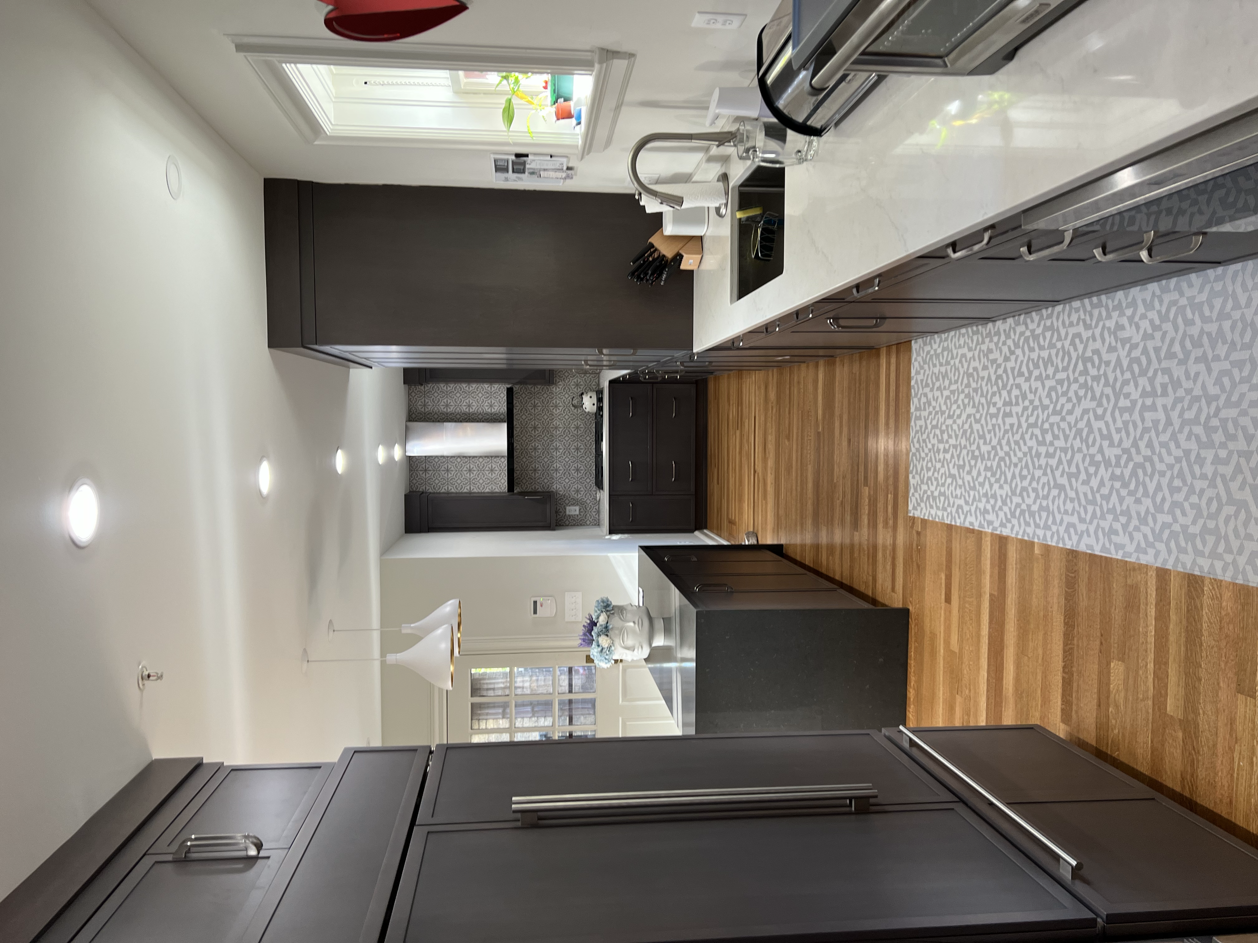 A modern kitchen with dark cabinets, white countertops, stainless steel appliances, a window with potted plants, and a patterned gray and white runner rug on a wooden floor.