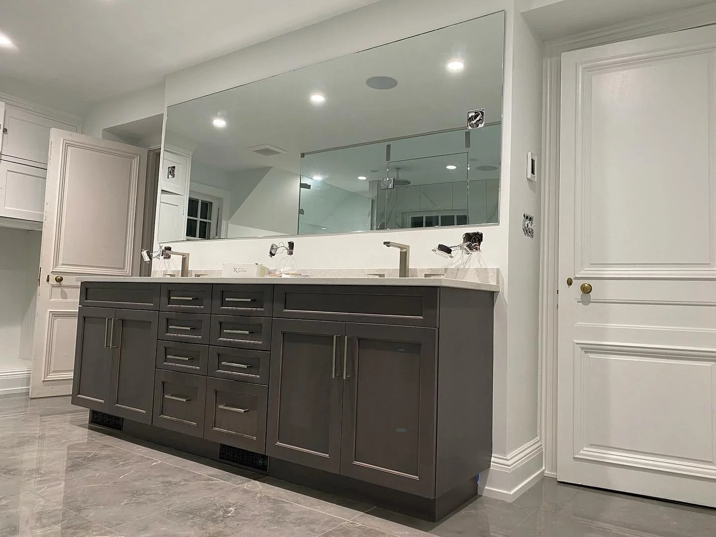 Bathroom vanity with dark gray cabinets, white marble countertop, reflected in large wall mirror, with two sinks and modern fixtures, in a white-walled room under construction.