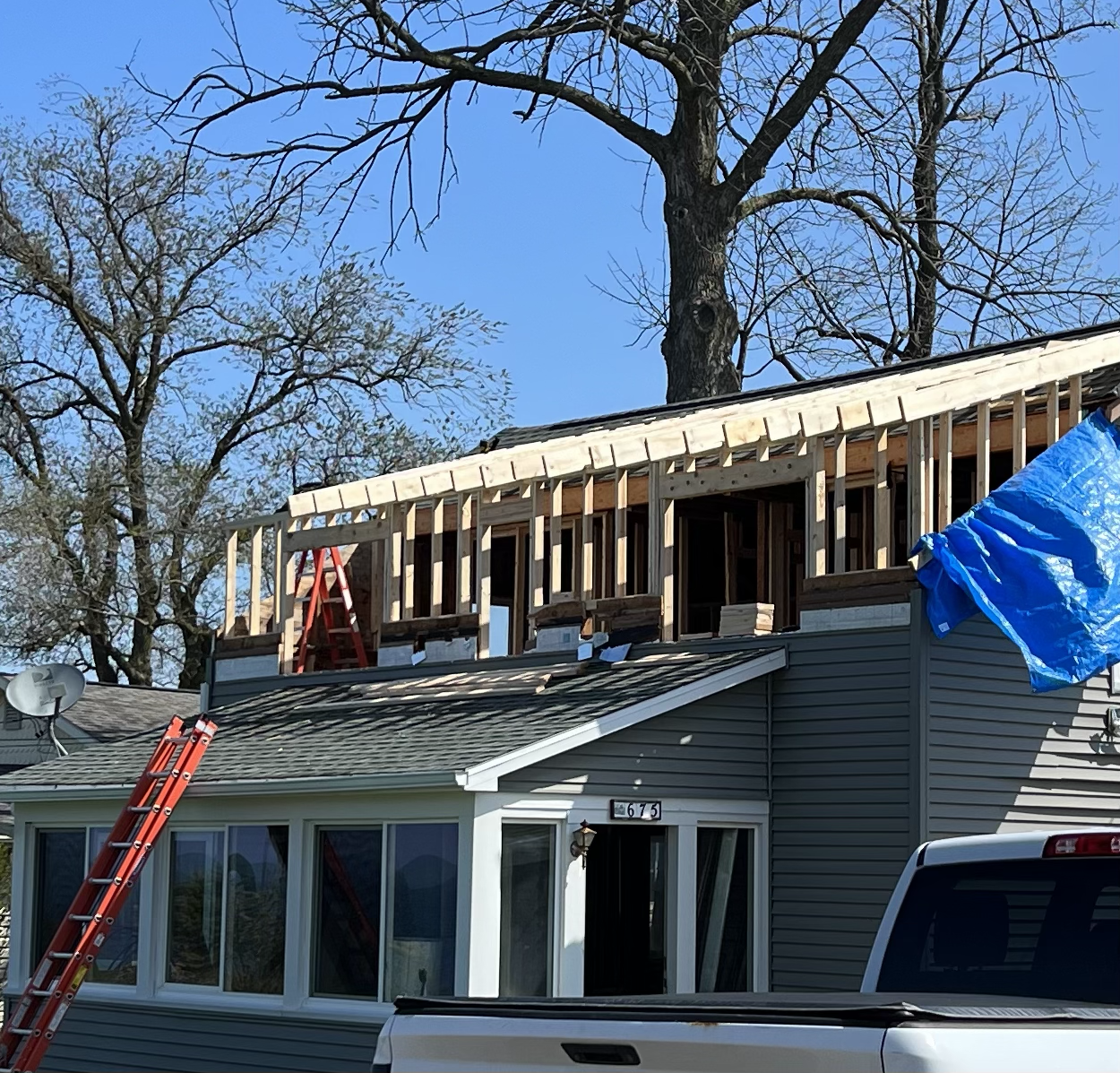 A house under construction with an added second-story frame. A red ladder leans against the house, and another red ladder is positioned on the roof. Blue tarp covers part of the construction area. The house has a grey exterior with a bay window at th