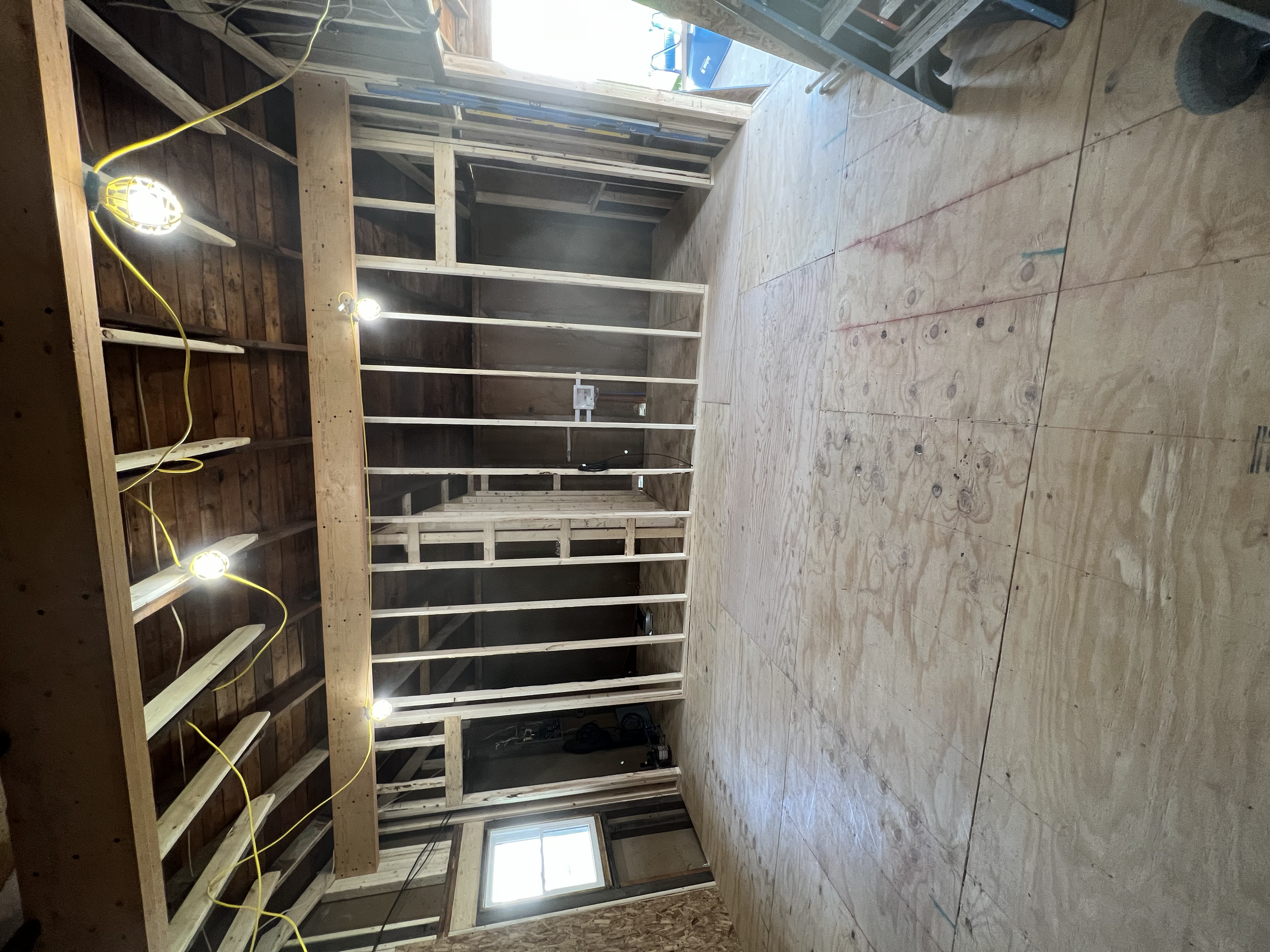 Interior view of a building under construction with exposed wooden framing, plywood flooring, and temporary lighting.