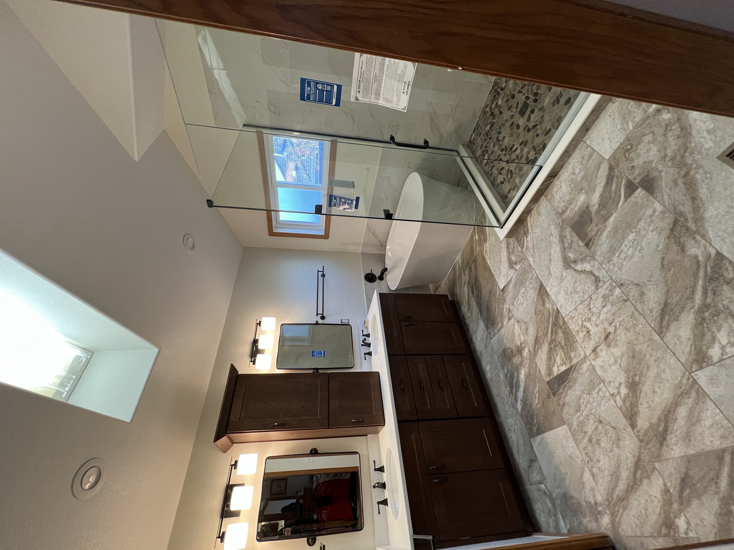 A modern bathroom featuring a glass shower enclosure, a freestanding bathtub, a double vanity with dark wood cabinets, large framed mirrors, wall-mounted lights, and a skylight providing natural light, with marble and tile flooring.