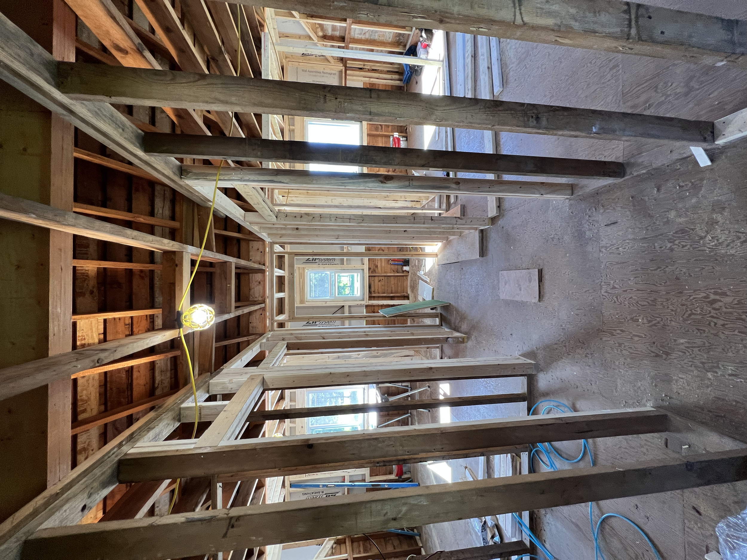 Interior of a house under construction with exposed wooden framing, several windows installed, and a temporary light fixture hanging from the ceiling.