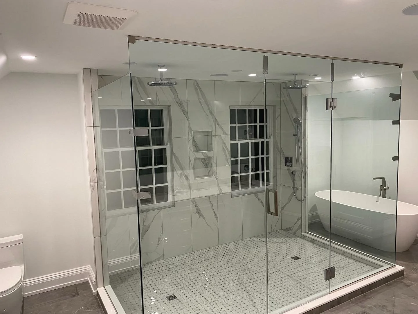Modern bathroom with a large glass-enclosed shower featuring white marble walls, tiled floor, and built-in shelves, a freestanding bathtub, and a window with grid panes.