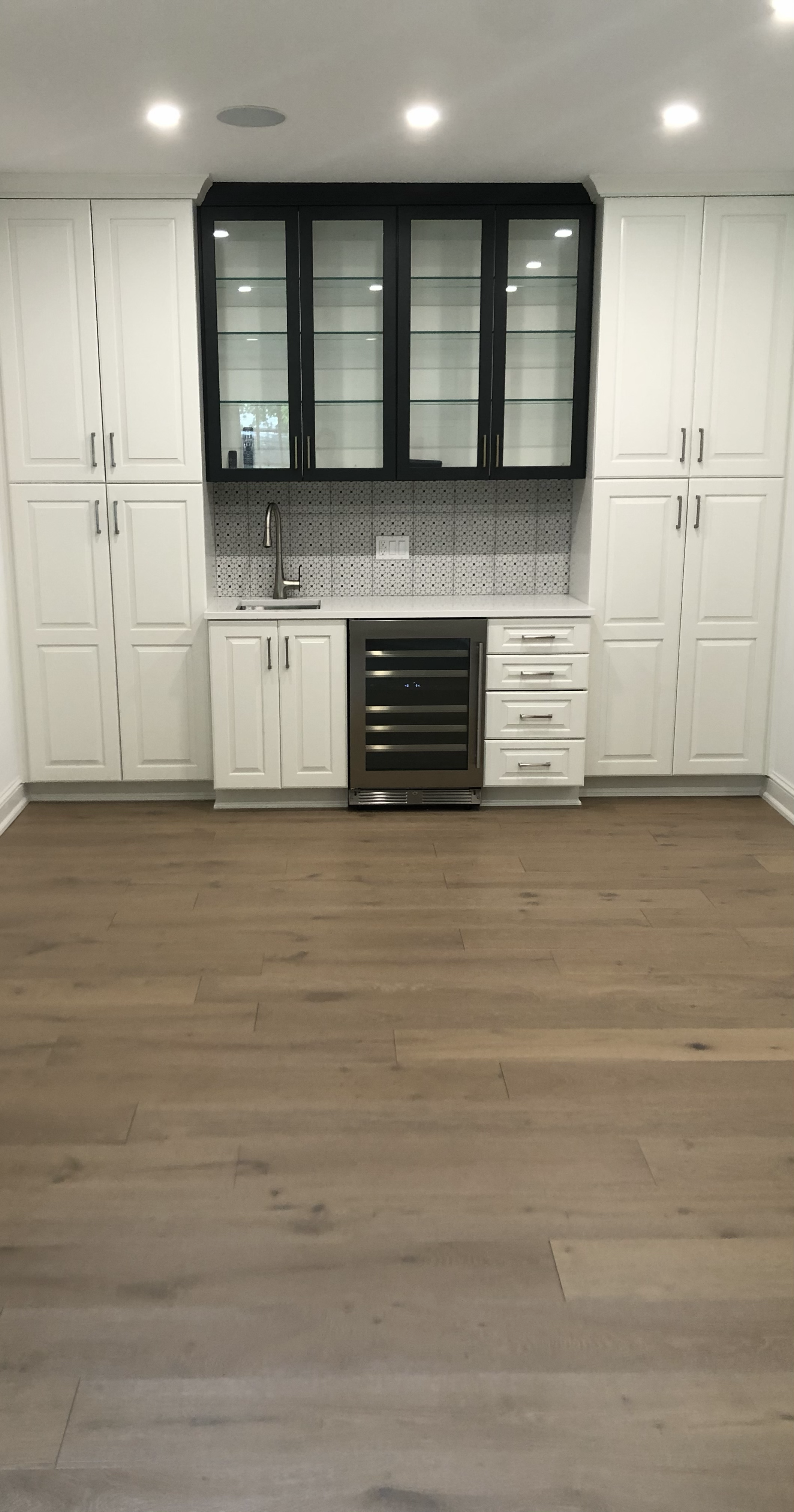 Kitchen with white cabinets, black upper cabinet with glass doors, stainless steel wine cooler, small faucet, tiled backsplash, and wooden floor.