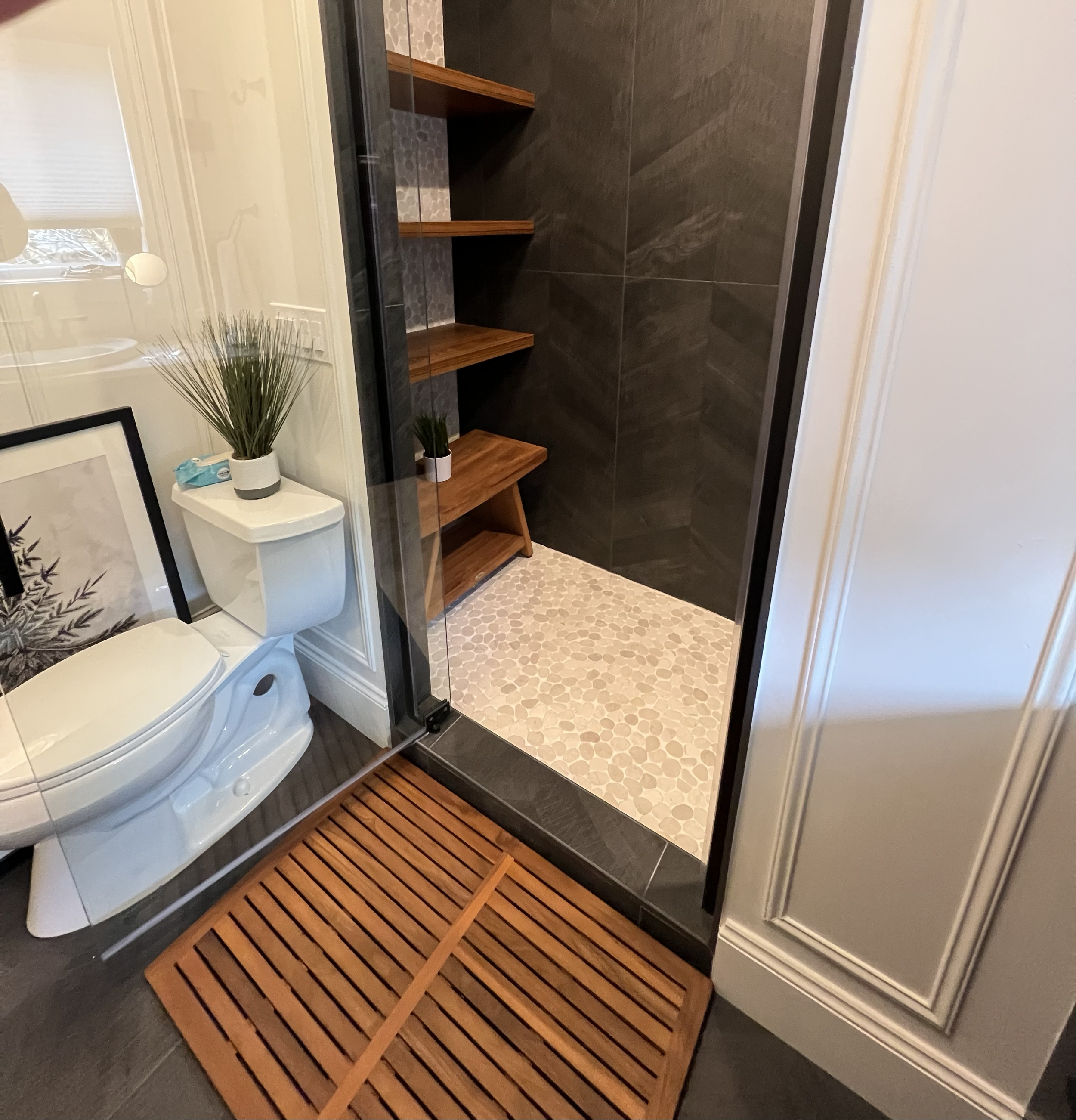 Bathroom with dark tiled walls and pebble tile floor. Wooden shower bench and slatted wooden mat outside shower. White toilet and small plants on nearby shelf.