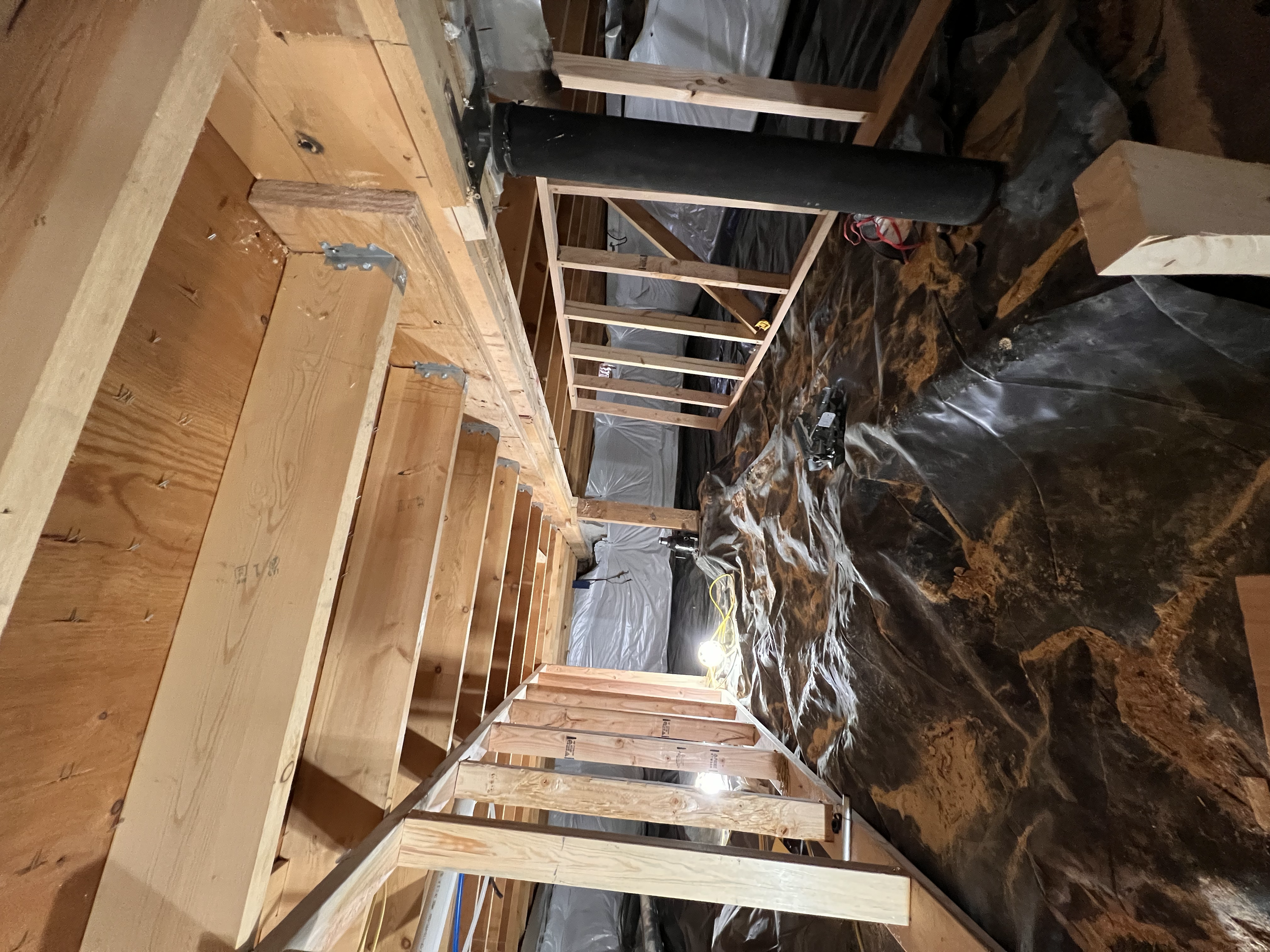 Construction site with wooden framing and black pipe, plastic sheeting covering unfinished floor, work light on floor, tools, and materials scattered around.