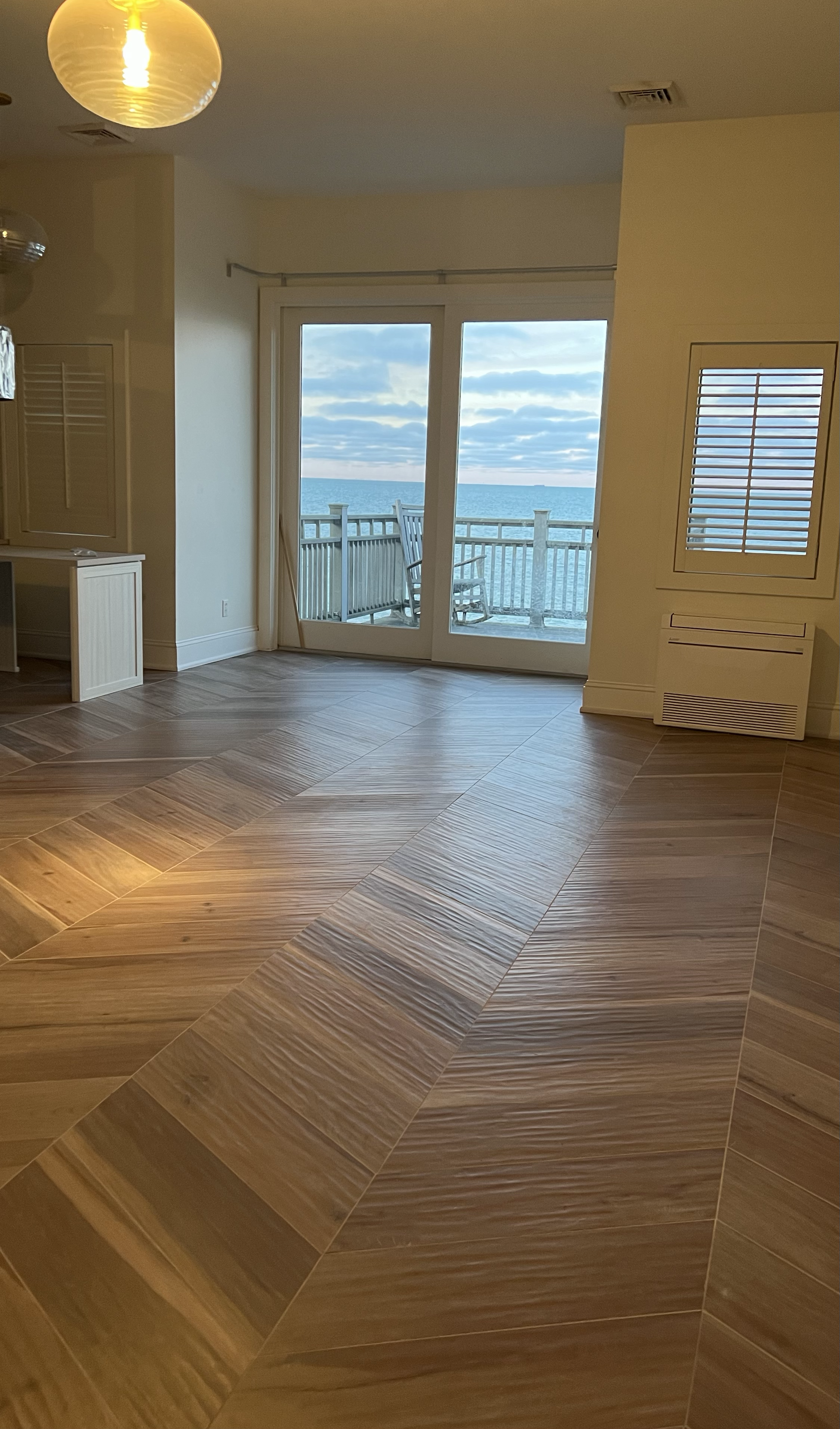 Room with wooden tile flooring and large sliding glass doors leading to a balcony overlooking the ocean, with a rocking chair on the balcony.