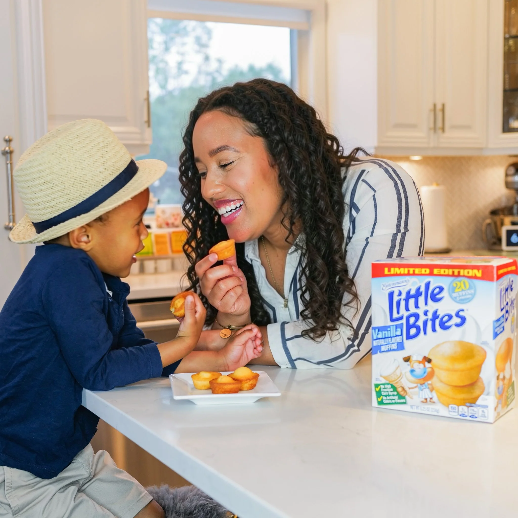 A young boy and woman are sharing a snack together at a kitchen counter, with a box of Little Bites vanilla muffins nearby.