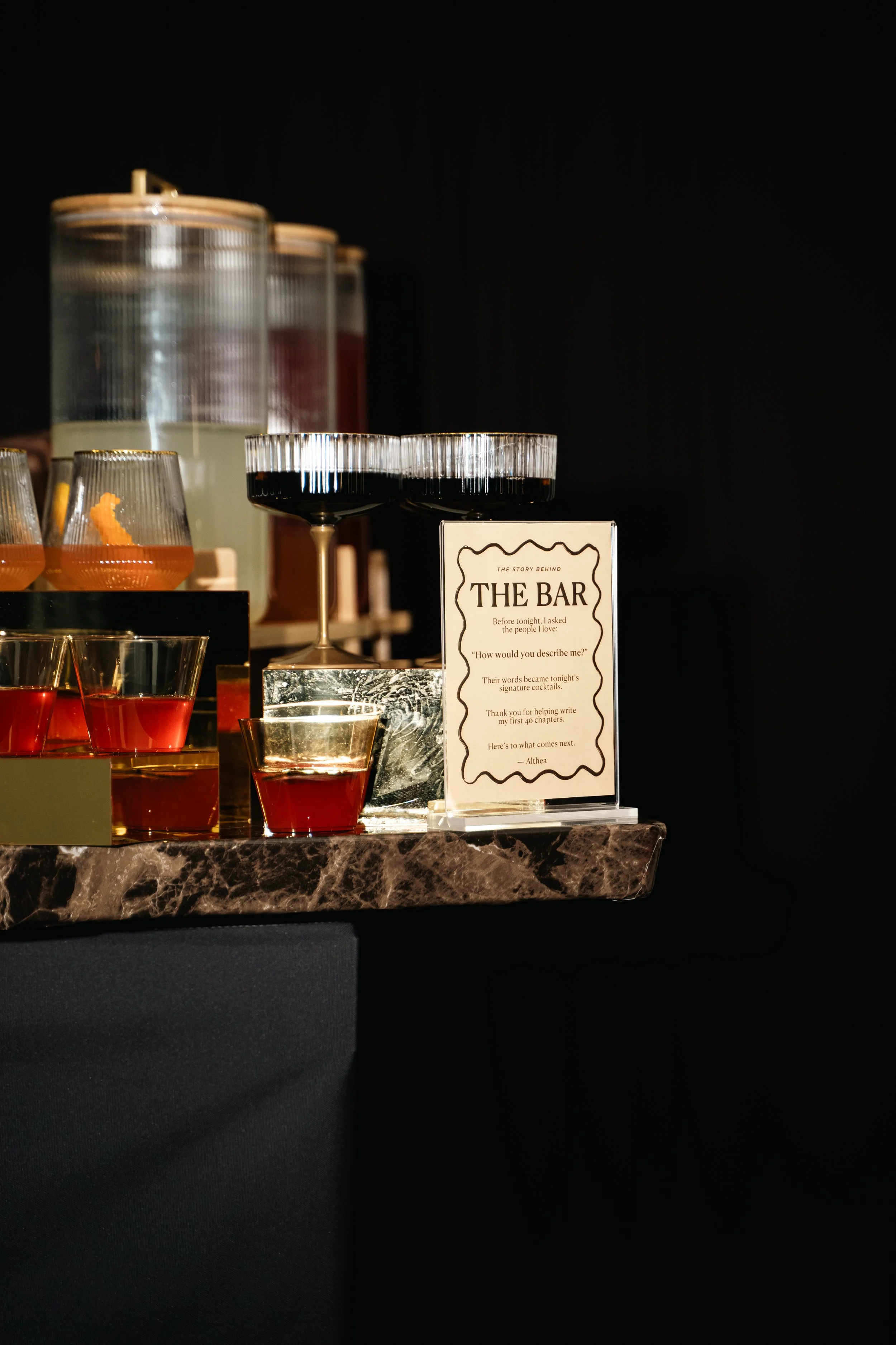 A display of various cocktails on a bar counter with a sign titled 'The Bar' that describes the story behind the drinks.