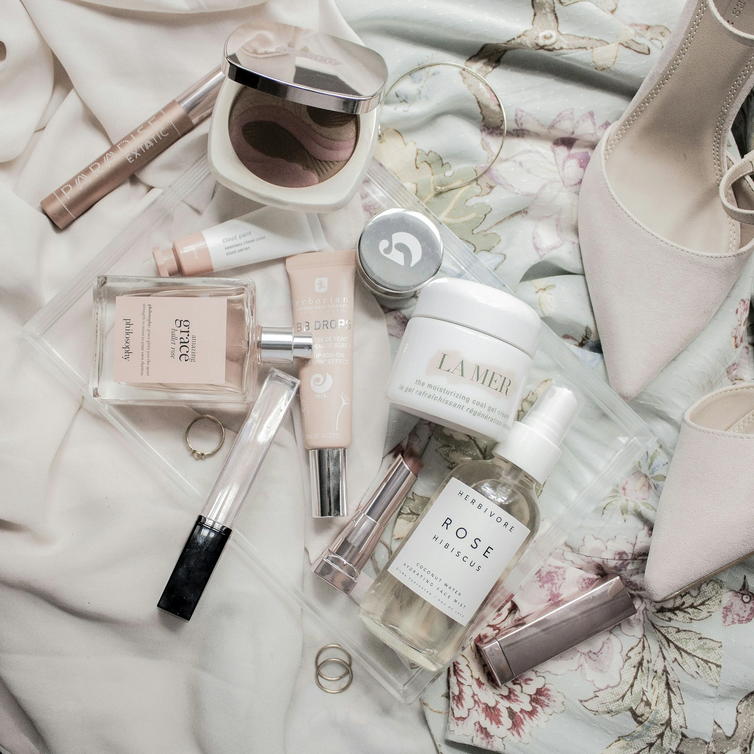 A collection of beauty and skincare products including a compact mirror, foundation or concealer, moisturizers, and a perfume bottle arranged on a soft surface with floral fabric in the background.
