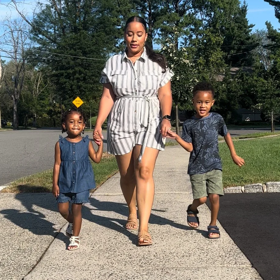 A woman walking on a sidewalk holding the hands of two children, one girl and one boy, on a sunny day with trees and a street in the background.