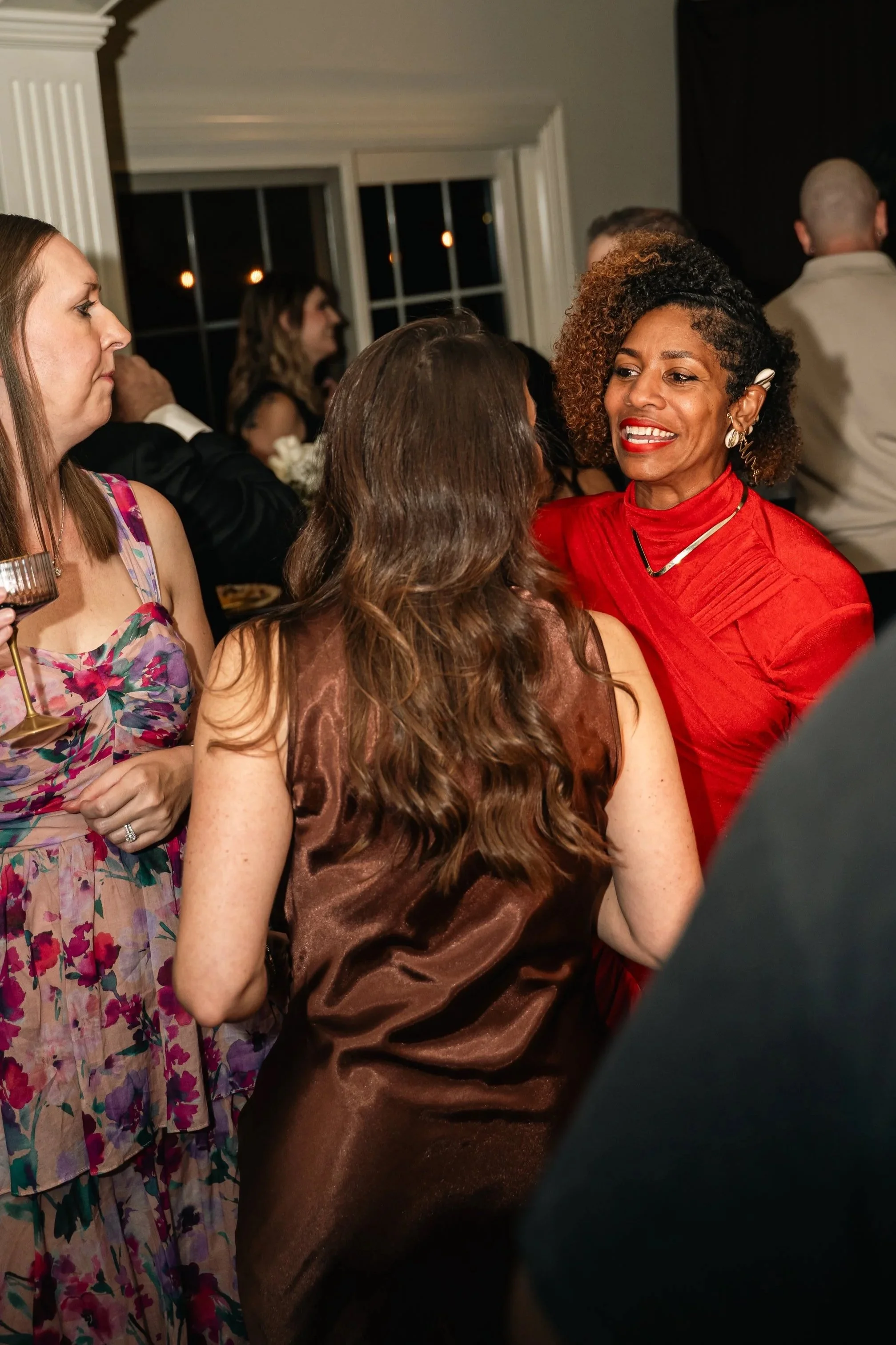 Smiling woman in a red dress engaging in a conversation at a social gathering, with other people in the background.
