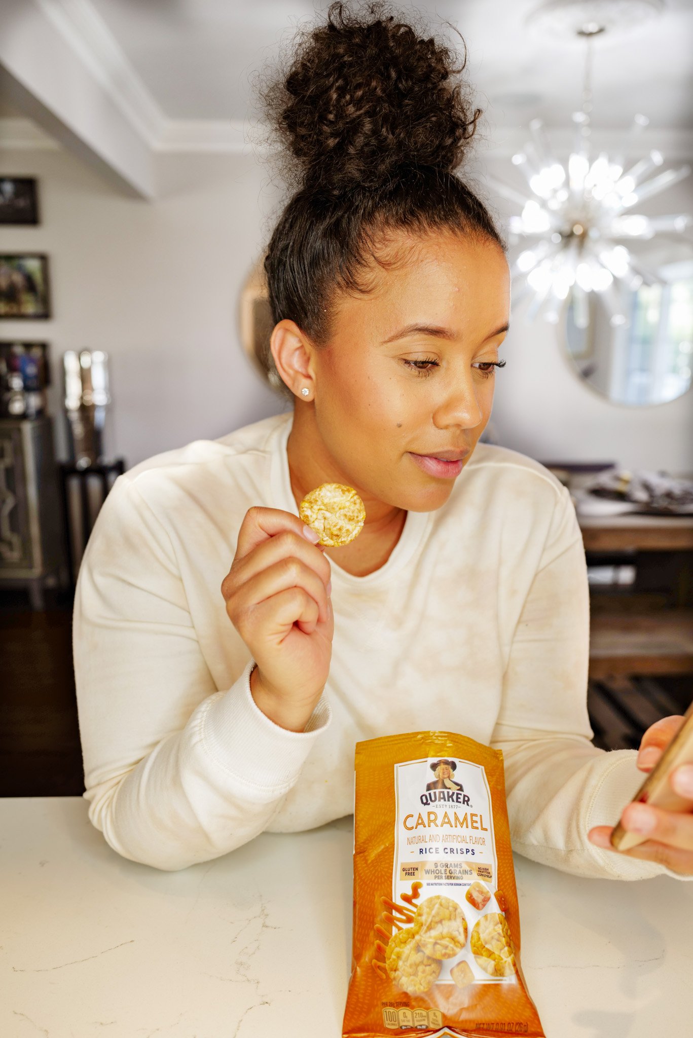 A woman with curly hair tied up in a bun is sitting at a table, holding a rice crispy treat in her right hand, with a bag of Quaker caramel rice crisps in front of her. She is looking at her phone.