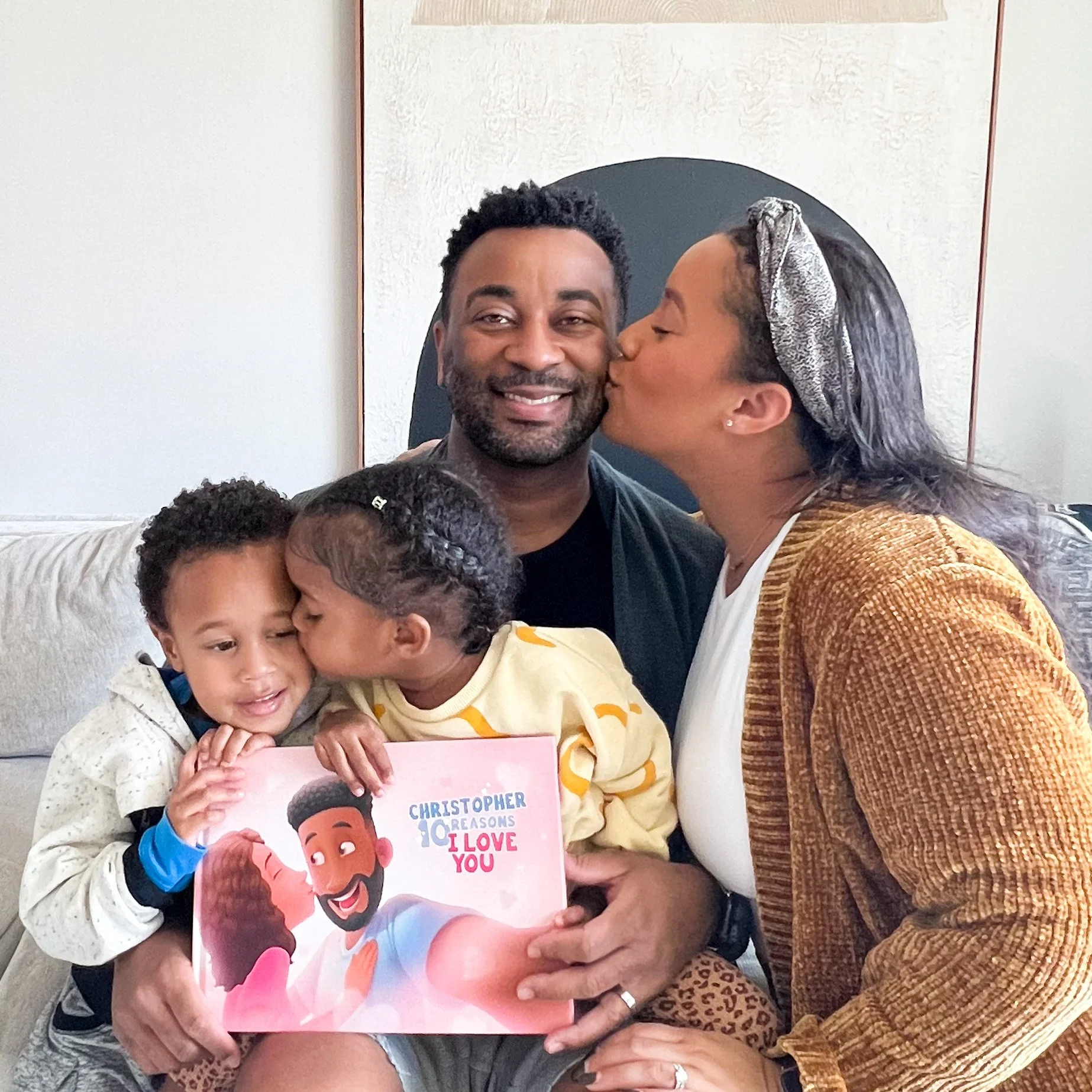 A woman is kissing a man on the cheek while holding an illustrated children's book titled 'Christopher Loves You'. There are children in the foreground, with one girl hugging the book. The background shows a white wall and framed artwork.