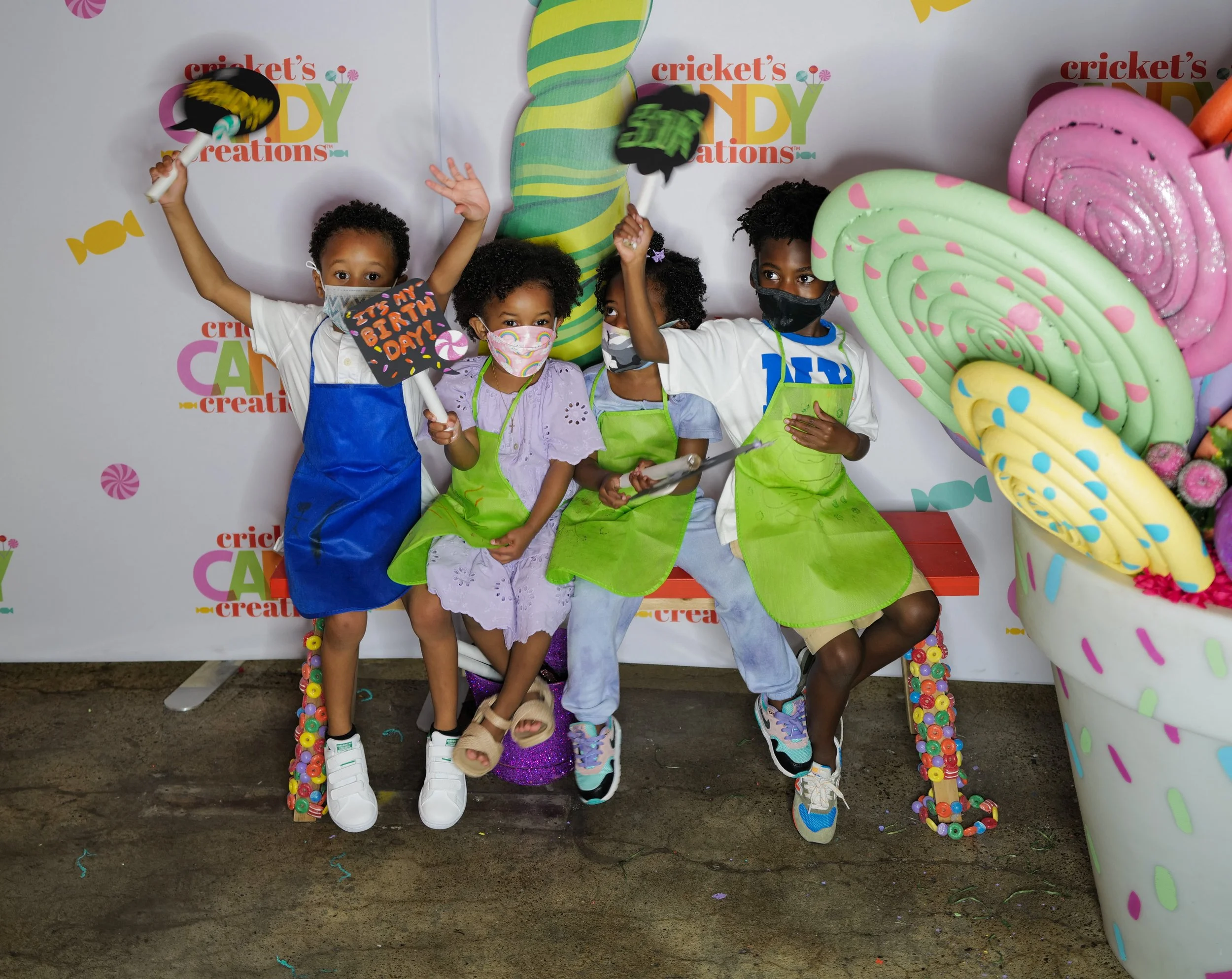 Children wearing face masks, sitting on a decorated chair, at a birthday celebration with large colorful fabric flowers and a backdrop reading 'Cricket's Candy Creations.'