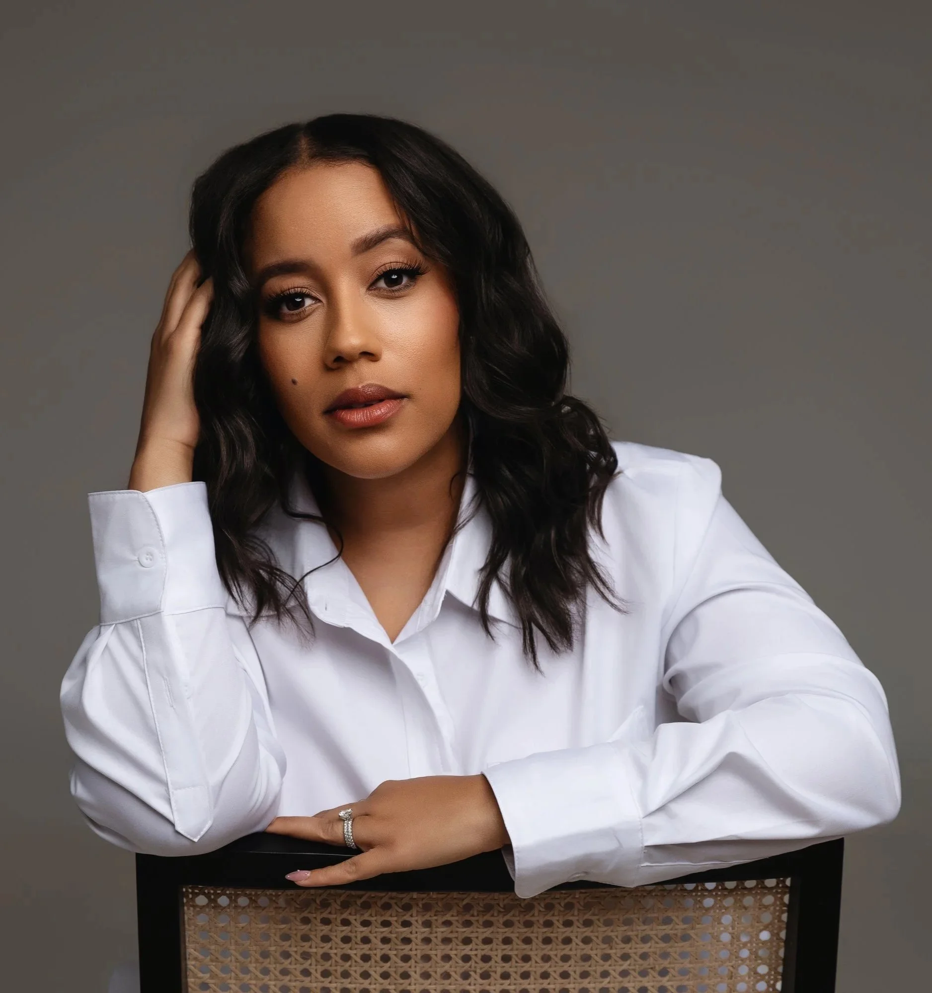 A woman with curly black hair and medium skin tone, wearing a white button-up shirt, is posing with her left hand resting on her temple. She is sitting at a table with a decorative woven panel, against a plain gray background.