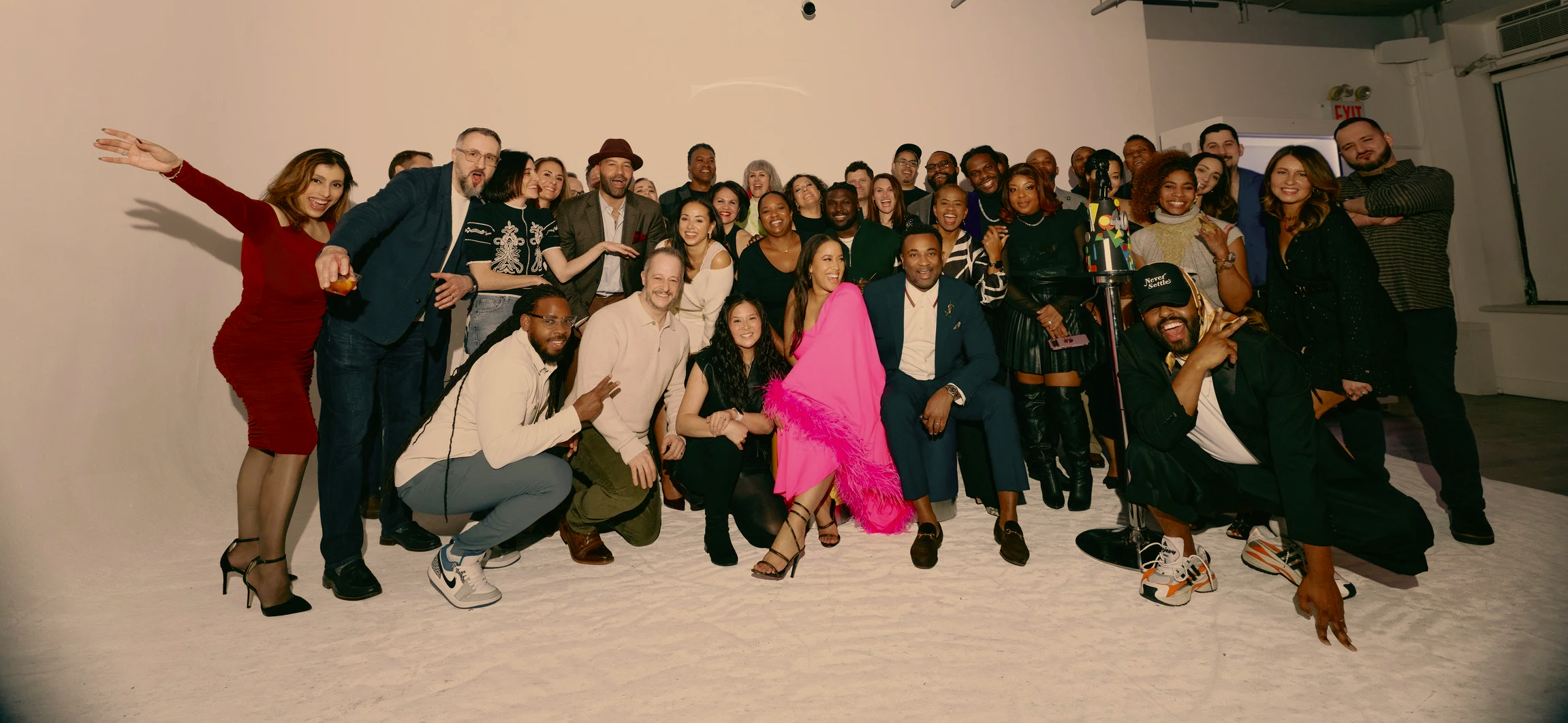 A large group of diverse, smiling people posing together in a photography studio, some waving, and others making gestures, with a plain white backdrop and some studio equipment visible.
