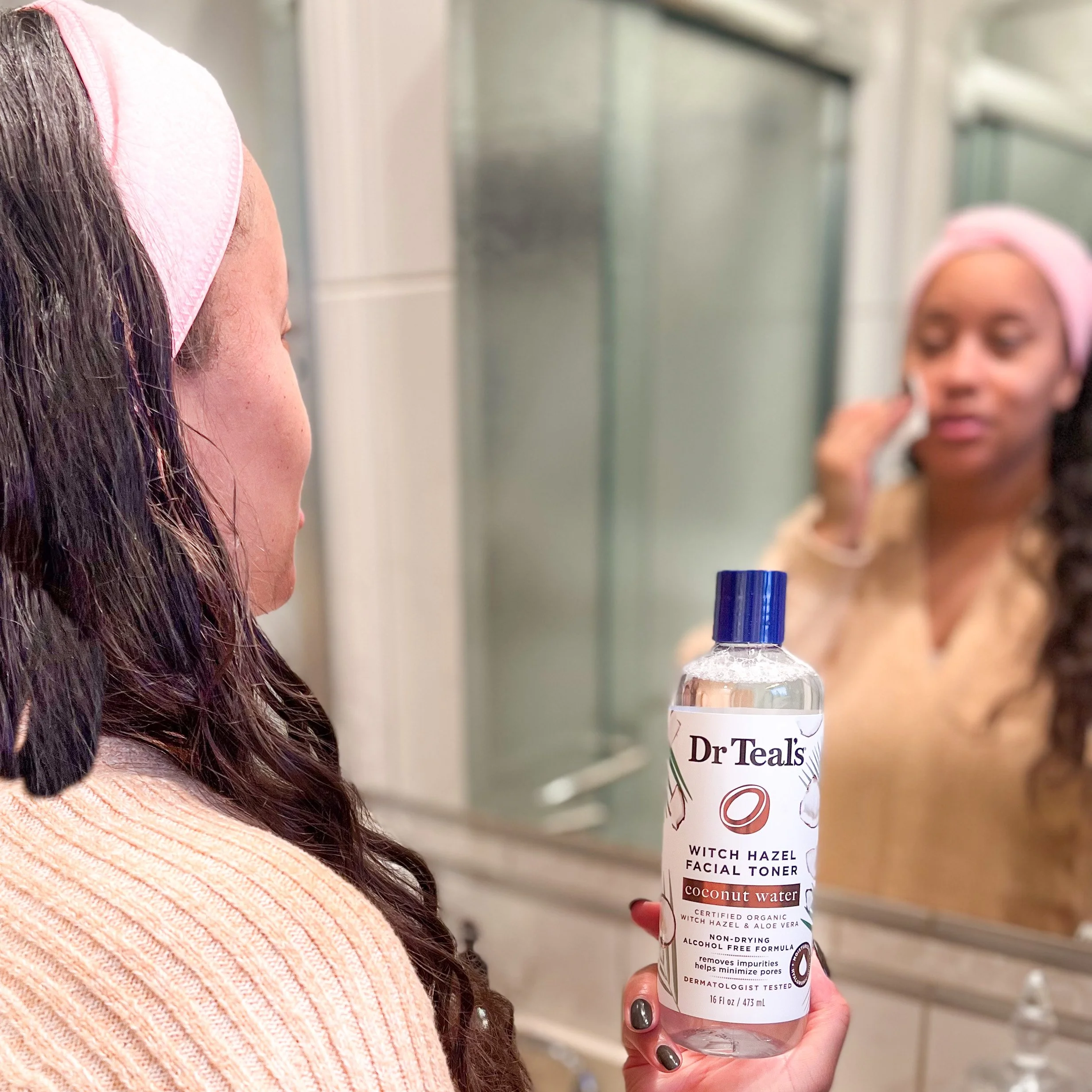 A woman in a light pink sweater holding a Dr. Teal's facial toner bottle in front of a mirror, with her reflection visible.