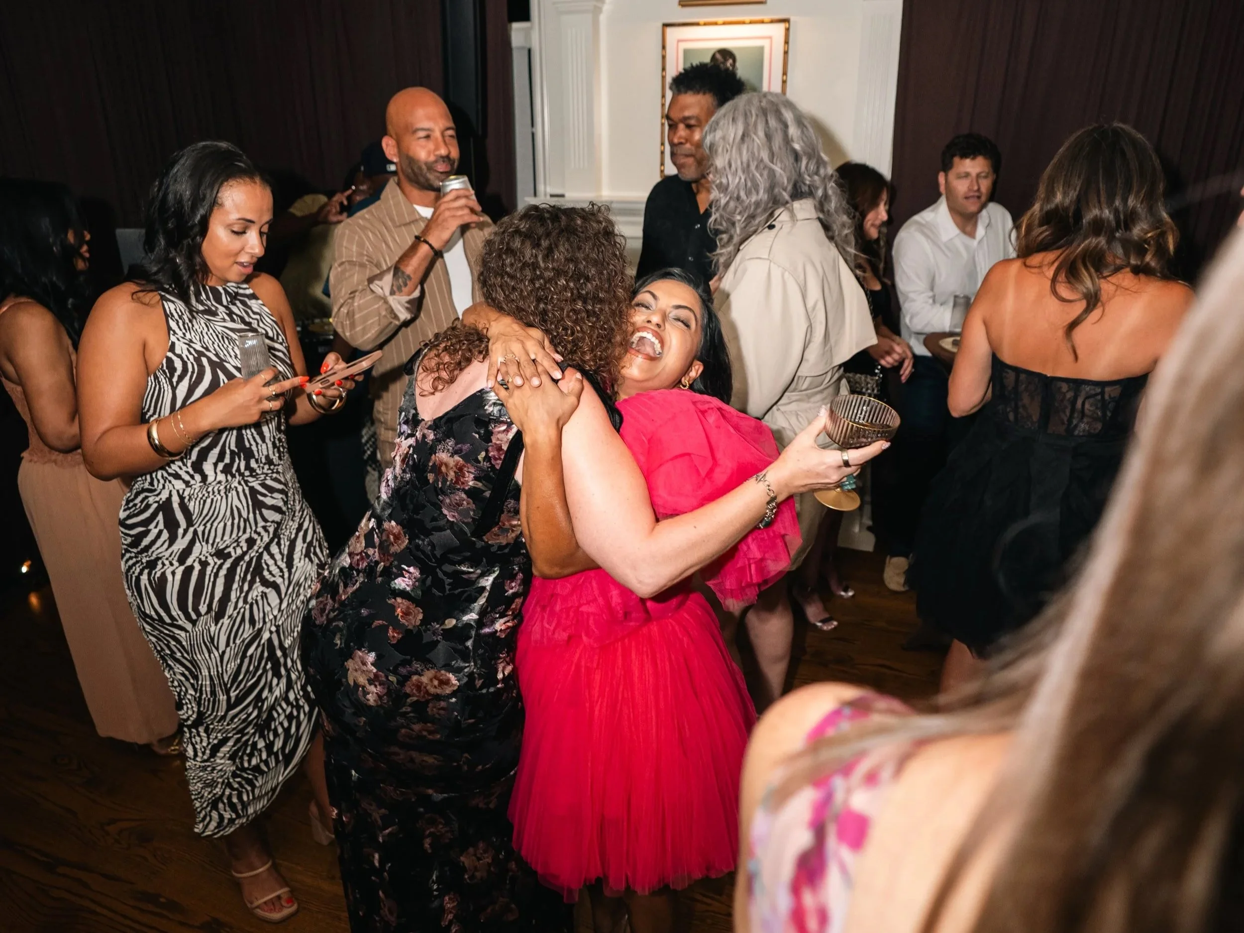 A diverse group of people are dancing and celebrating at a party, with some hugging, holding drinks, and socializing in a warmly lit room.