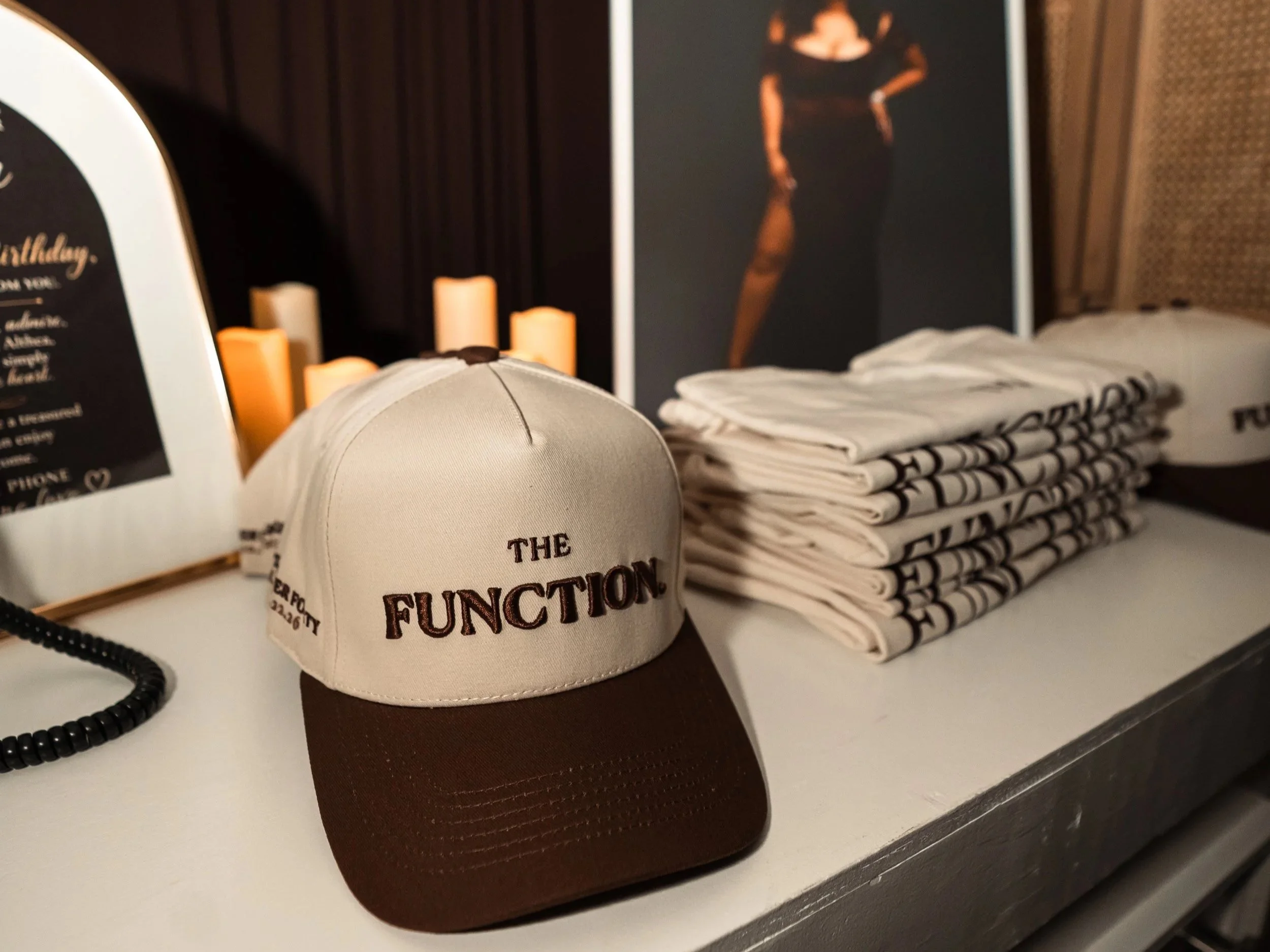 A beige hat with a brown brim and the text 'The Function' embroidered on it, placed on a shelf alongside folded white and black patterned towels, decorative candles, and a framed photograph of a woman in black clothing.