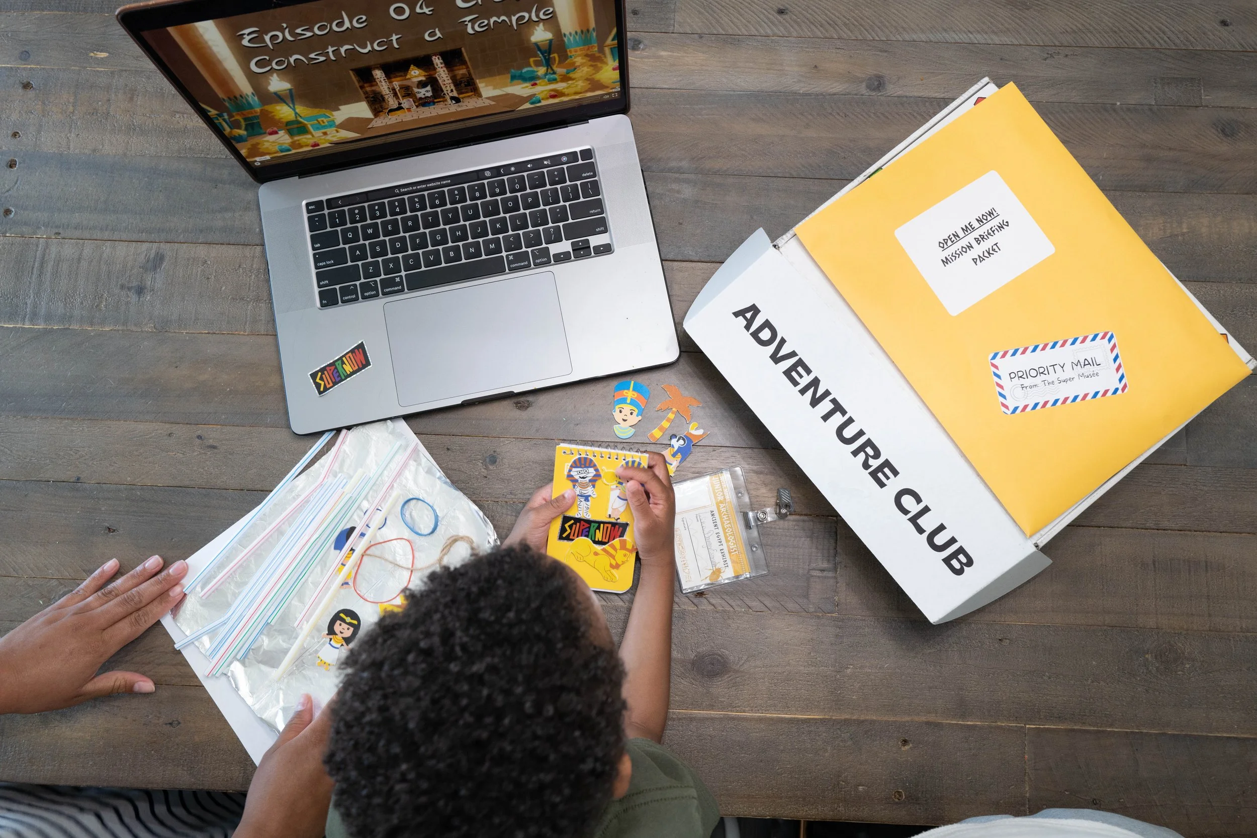 A child's hands holding a colorful sticker sheet featuring characters like a pharaoh, cat, and Egyptian figure. A person’s hand is visible on the left side, and a child's head is in the lower center. There are various supplies on a wooden surface, including a laptop, yellow folders labeled "Adventure Club," a clear folder with papers, and loose stickers. The laptop screen displays a website with the text "Episode 04: Construct a Temple."'}