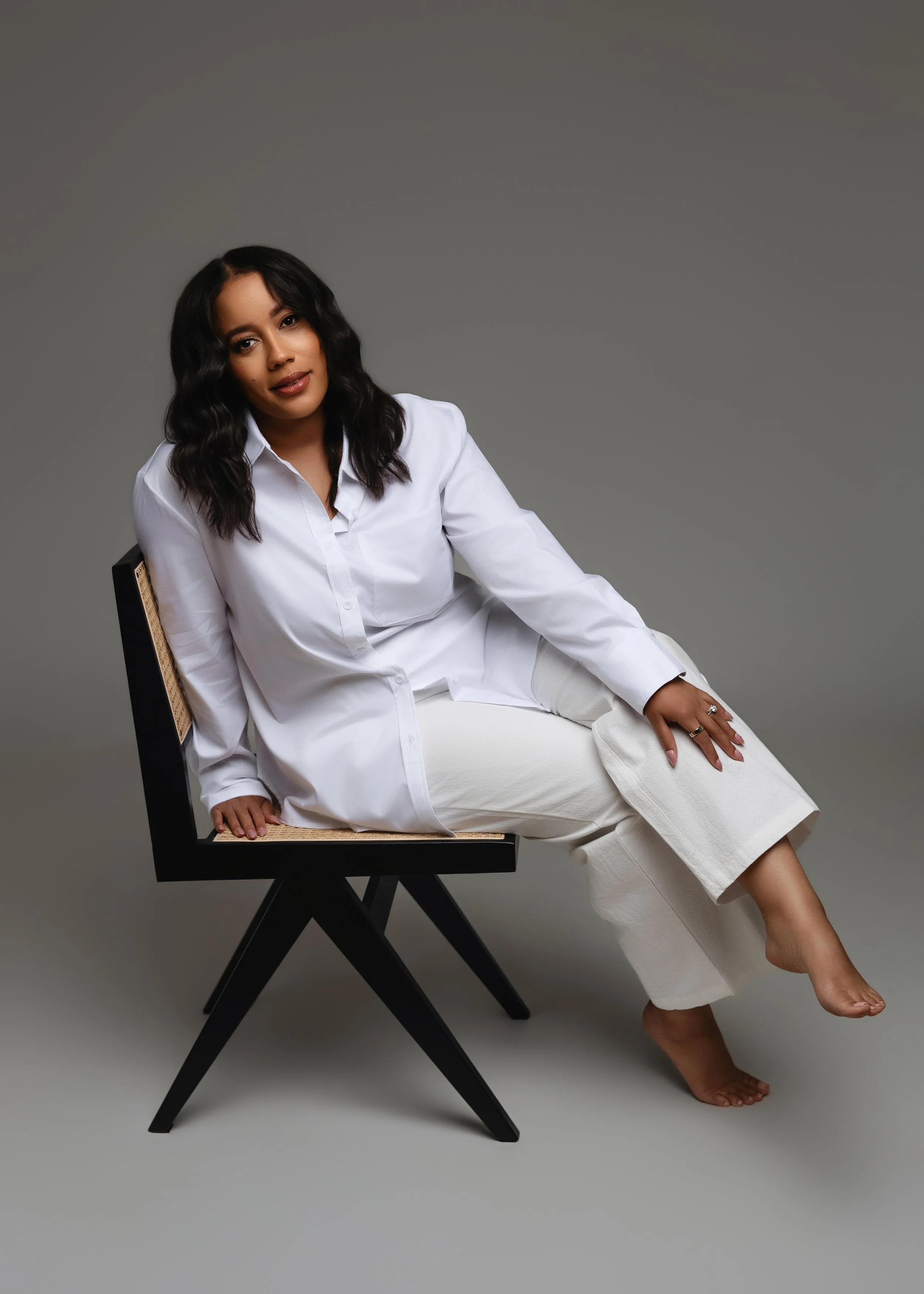 MsTrueHappiness. A woman with dark wavy hair wearing a white button-up shirt and white pants sitting on a black chair against a plain gray background.