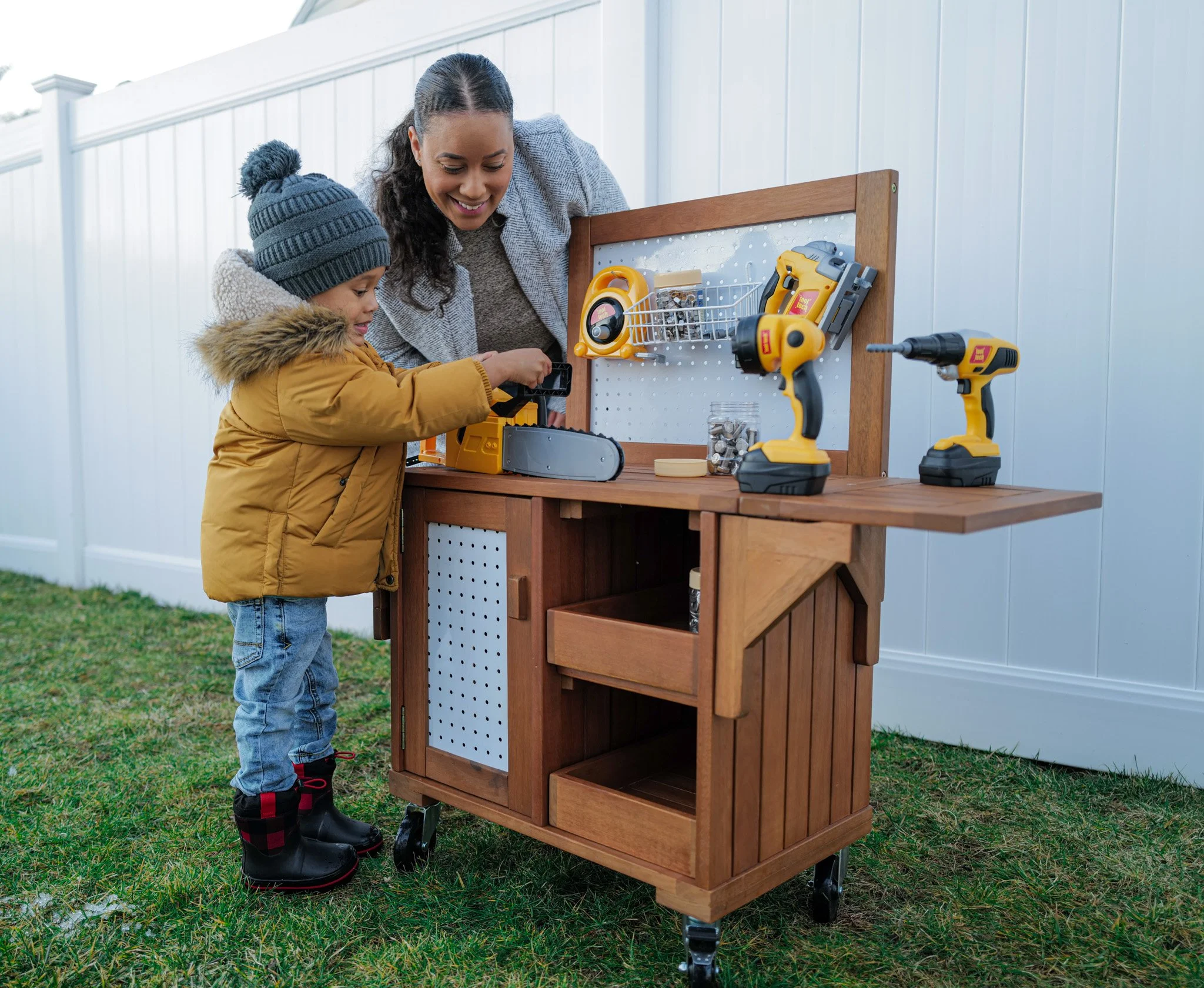 A woman and young boy with winter clothing, including a hat and boots, are setting up a wooden outdoor work station with power tools, a drill, and hand tools, on a grassy yard with a white fence.