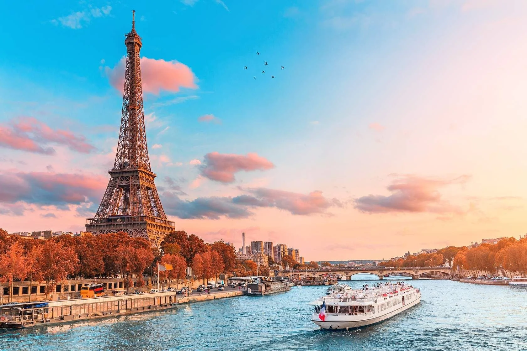 A cityscape featuring the Eiffel Tower beside the Seine river at sunset, with orange and pink clouds, boats on the water, and tree-lined streets.