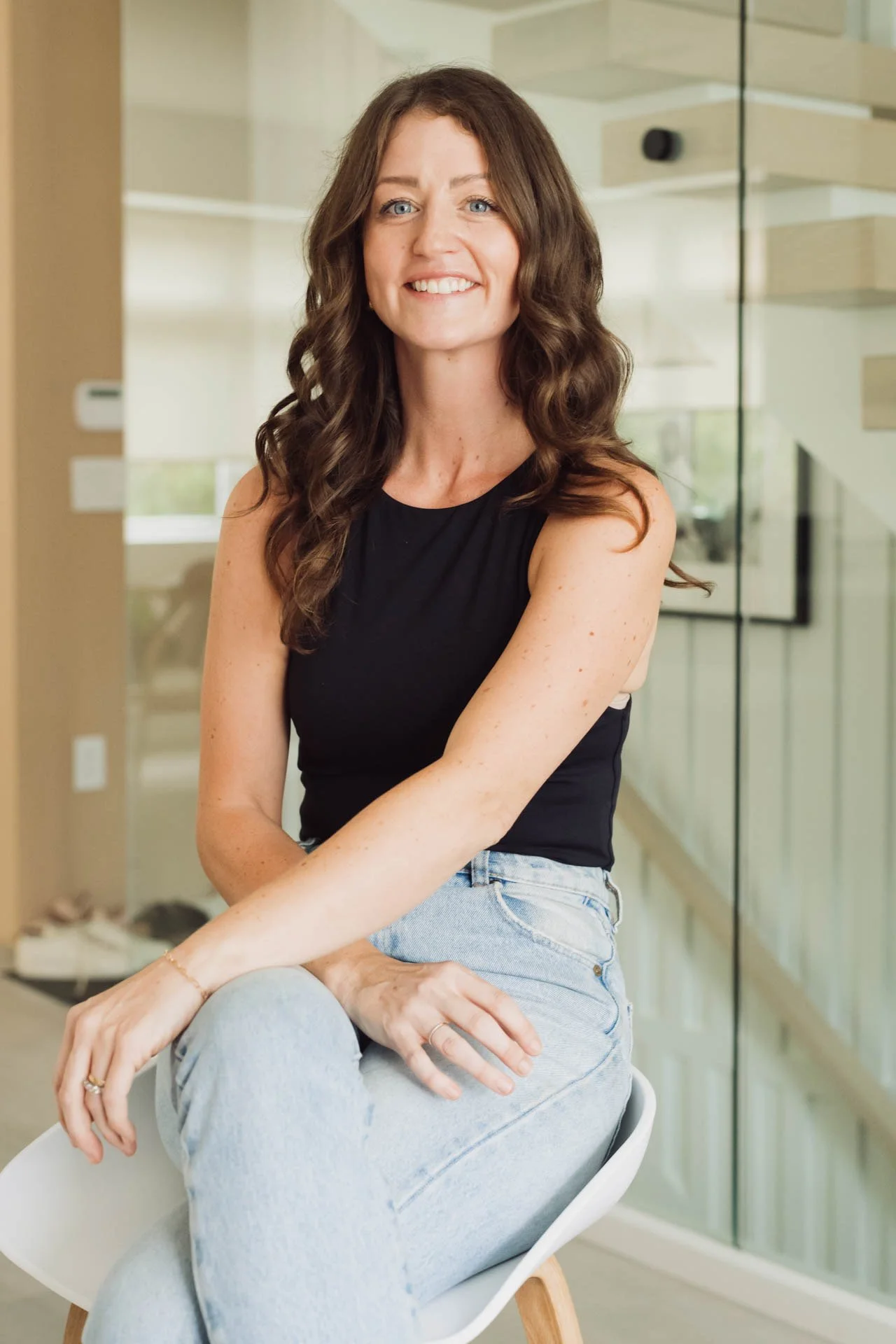 A woman with wavy brown hair, wearing a black sleeveless top and light blue jeans, sitting in a modern interior with a glass wall and stairs in the background.