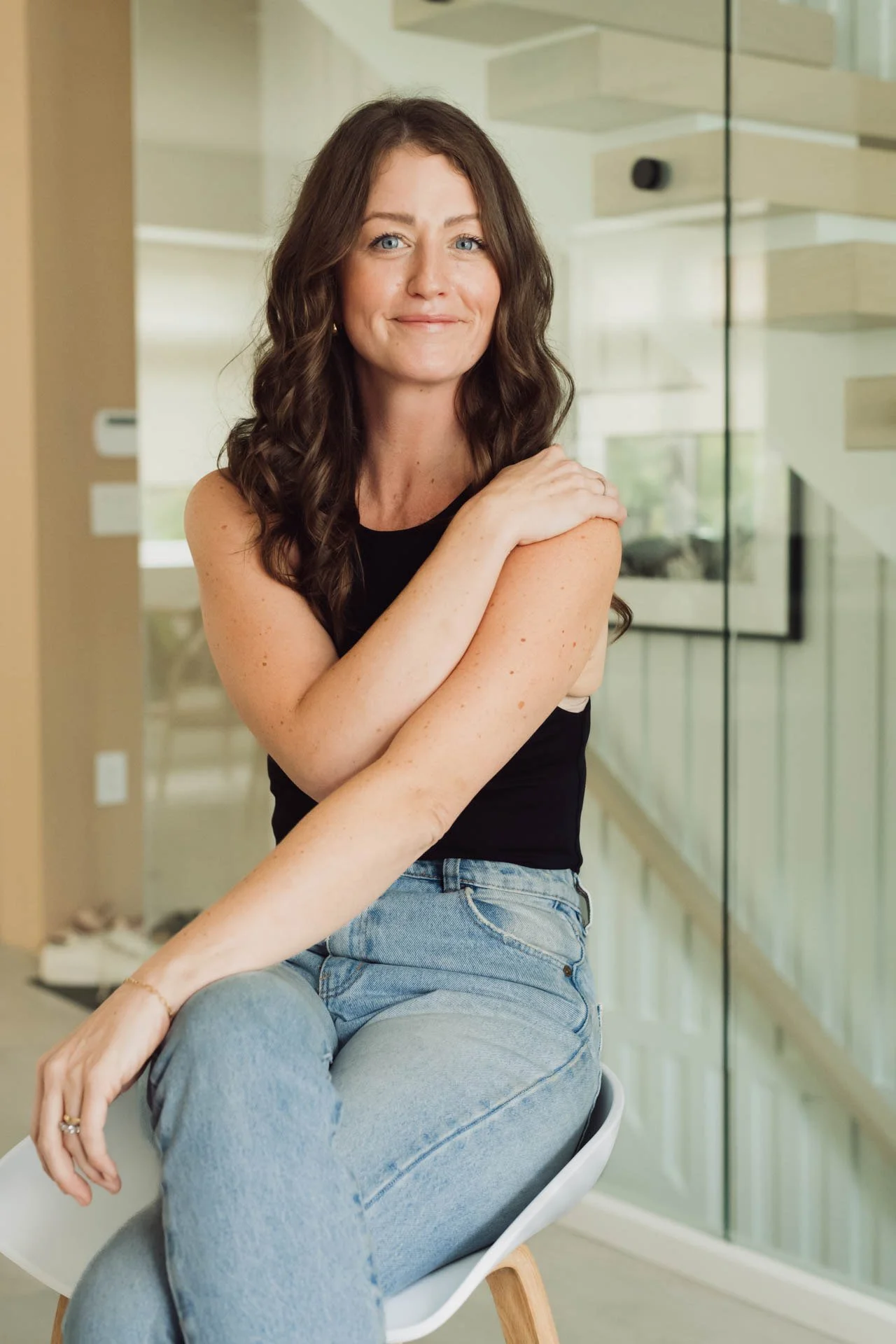 A woman with long wavy brown hair and blue eyes smiling while sitting on a white chair in a modern indoor space with glass walls.