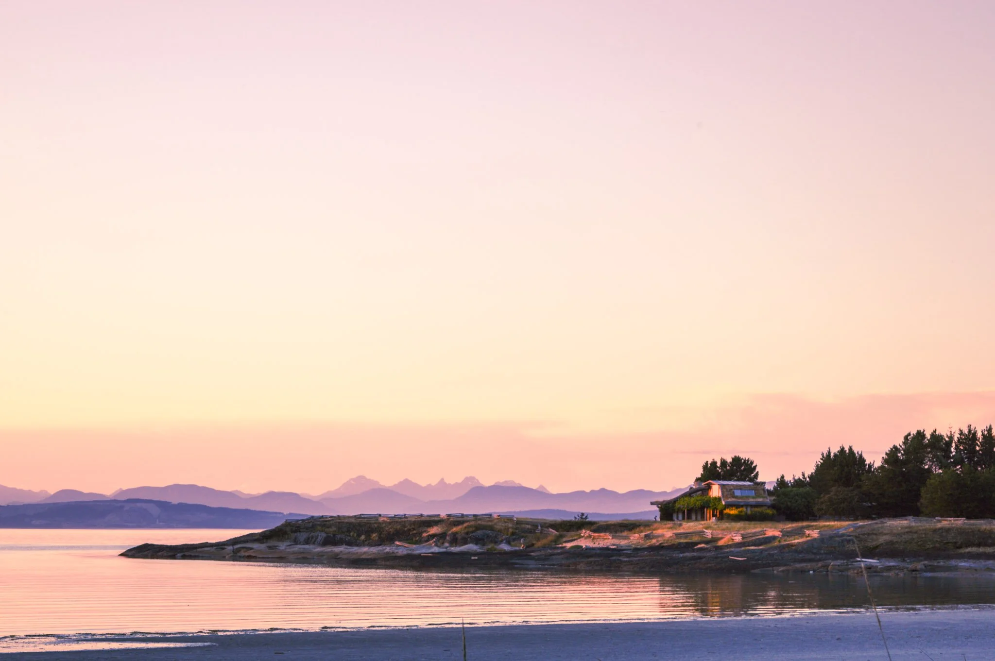 A peaceful lakeside scene at sunset with a pastel-colored sky, calm water reflecting the sky, a small house with solar panels, and distant mountains.