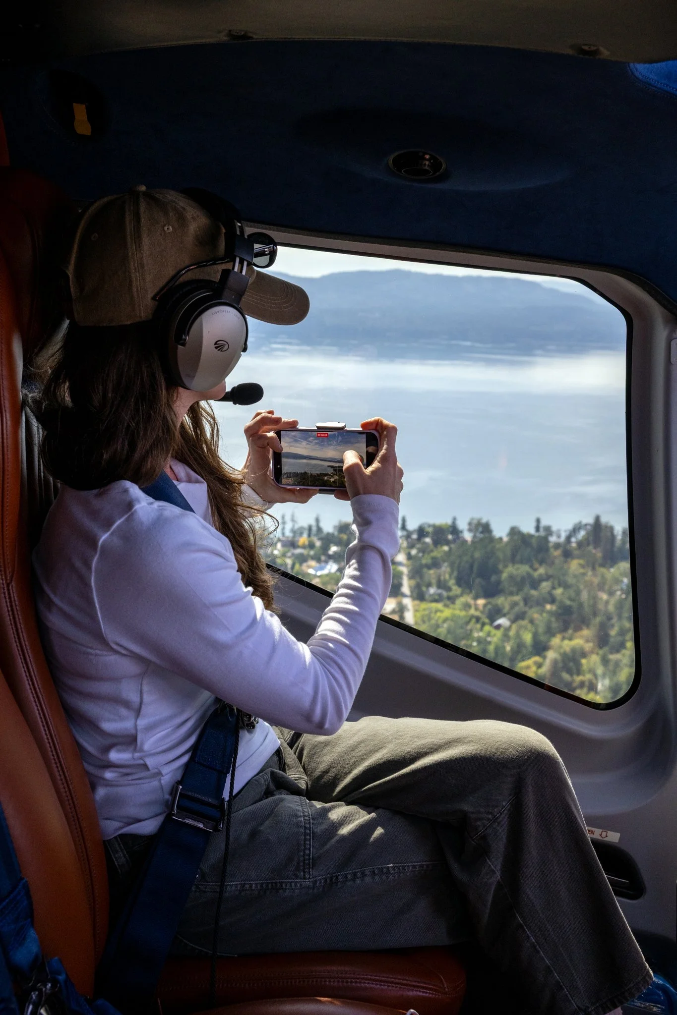 A woman with long, wavy hair wearing a beige cap and a white hoodie captures a photo through the window of a helicopter. She is seated on a brown leather seat, with a blue harness or strap across her lap. Outside the window, there is a scenic view of forested hills and a body of water.