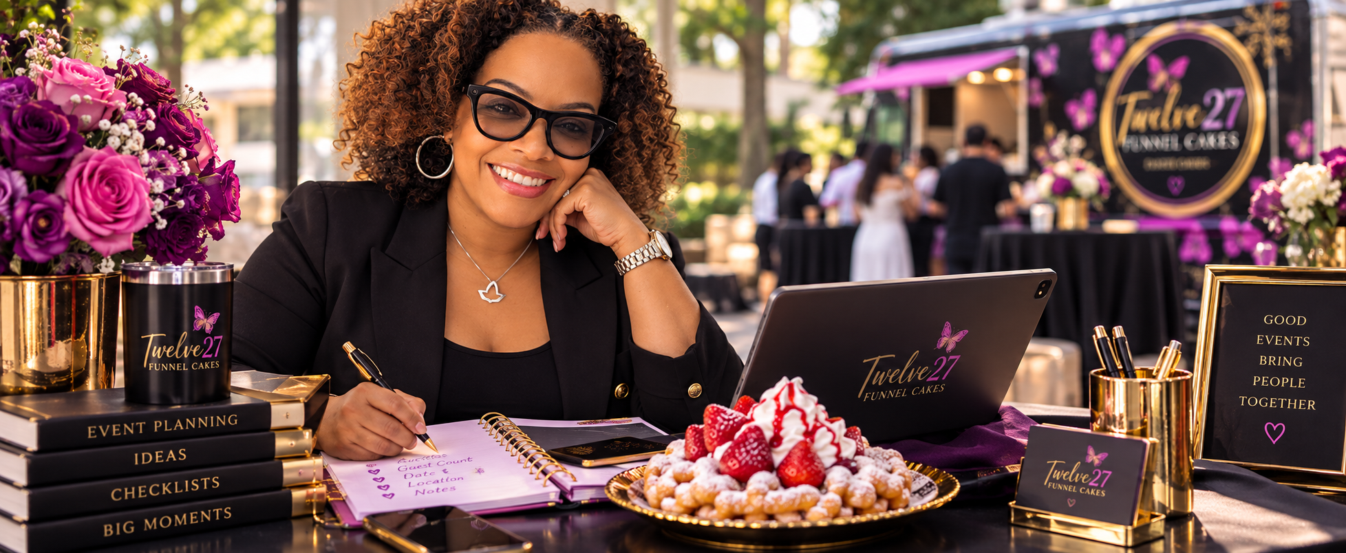 A woman with curly hair, glasses, and a black blazer sitting at a table decorated with purple and gold accents, including flowers and signage for Torelle 27 Funnel Cakes, smiling at the camera during an outdoor event. There are flower arrangements, a plate of strawberries with whipped cream, a laptop, and event planning books on the table.