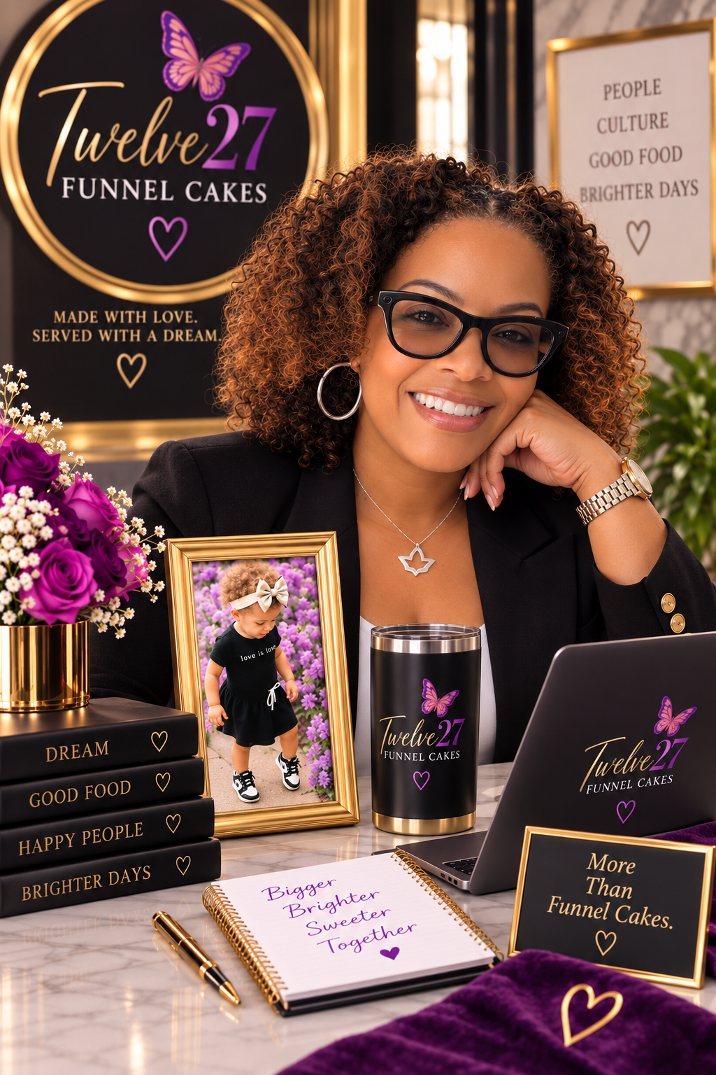 A woman with curly hair, glasses, and hoop earrings sitting at a table decorated with flowers, framed photos, and branded merchandise of Twelve 27 Funnel Cakes. There are signs on the wall behind her with inspirational phrases, and a notebook with a handwritten message reading 'Bigger, Brighter, Sweeter, Together'.