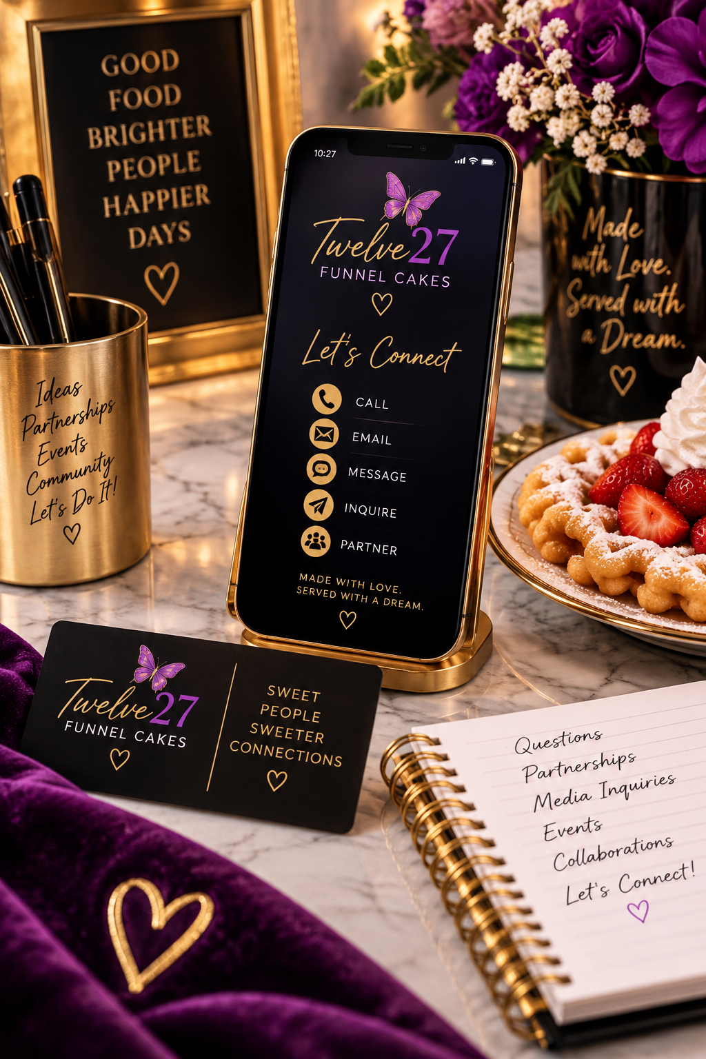 A smartphone screen displaying contact options for 'Twelve 27 Funnel Cakes' surrounded by floral arrangements, a plate of funnel cake with strawberries, a black card with purple butterfly and gold text, a gold pen holder with pens, a gold sign with motivational message, and a spiral notebook with handwritten contact prompts.