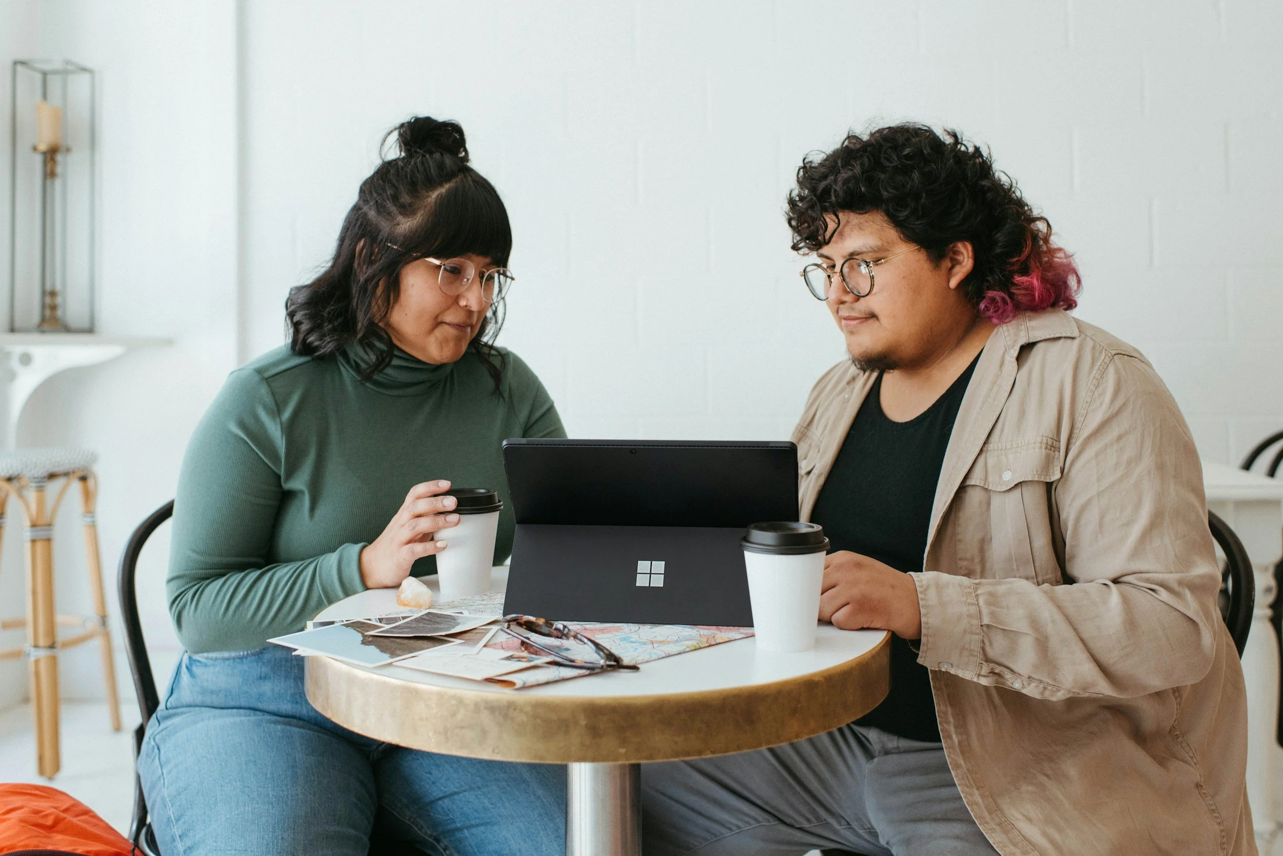 Two people with dark hair and glasses looking at a tablet or screen in conversation. Each person has a coffee cup.  One person has dyed pink curls and a beard.