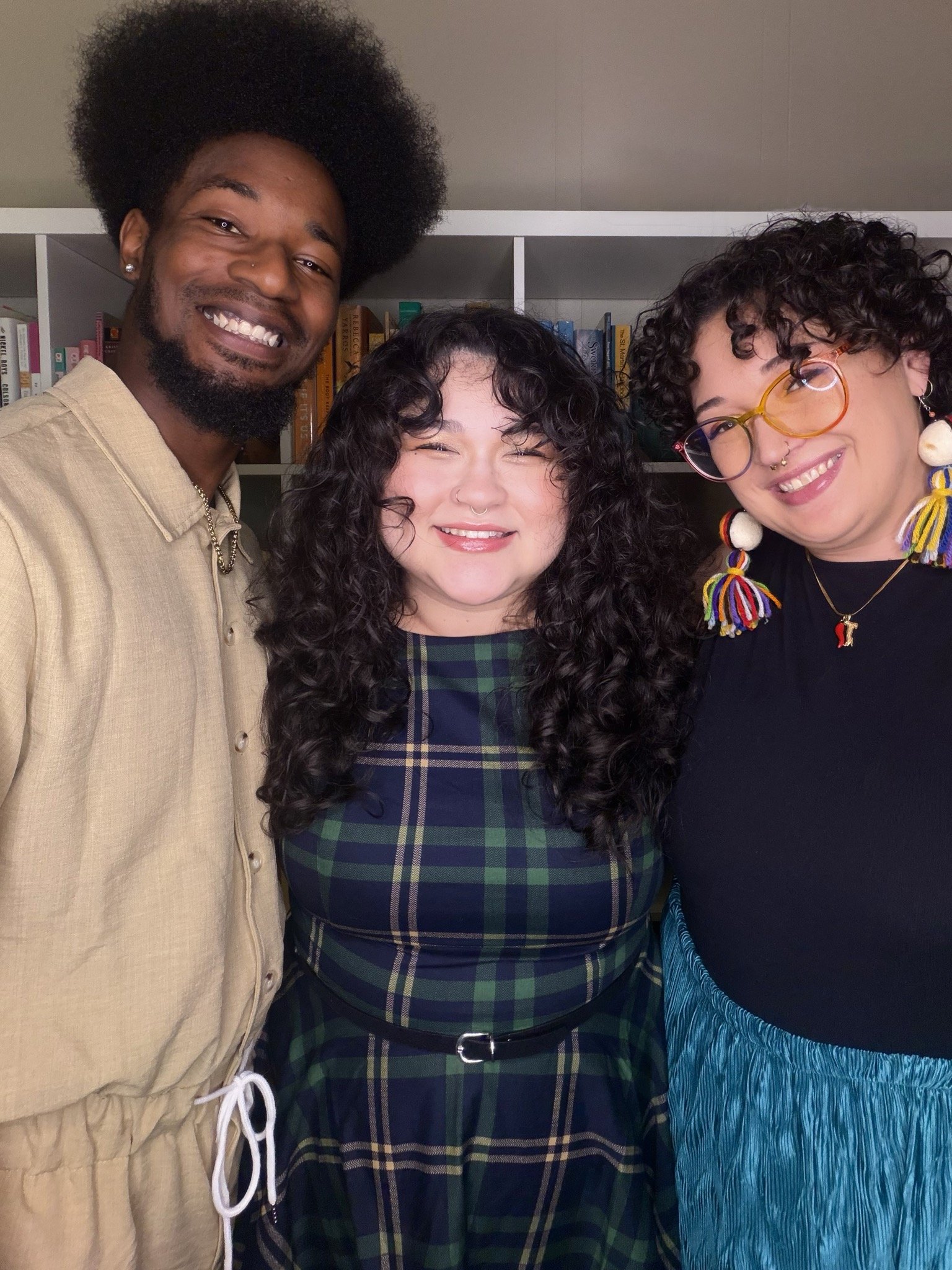 Three people smiling at the camera. Left person has an afro, while middle and far right have loose wavy hair. All three are leaning towards each other affectionately.